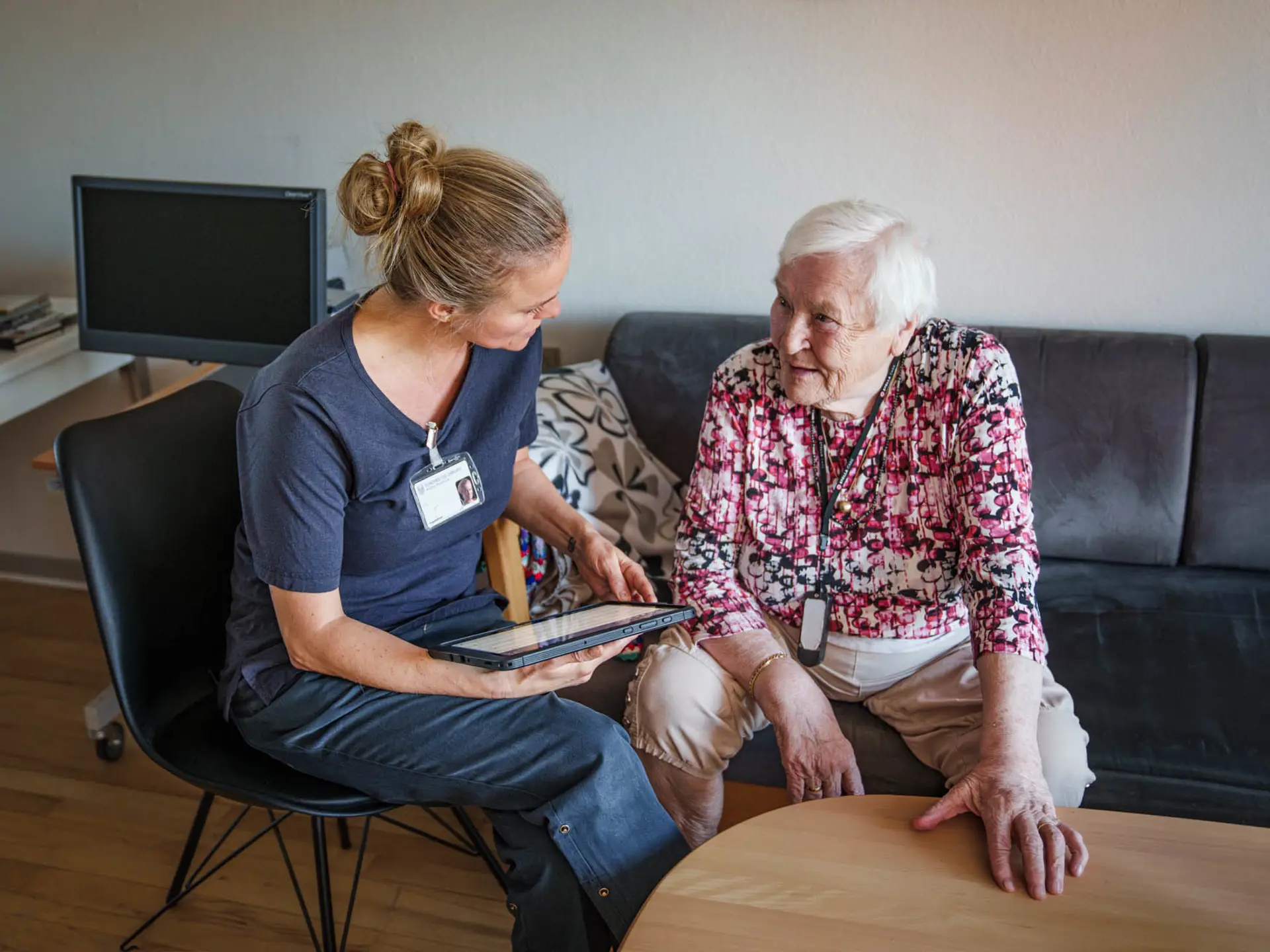 En plejepersonale viser en tablet til en ældre kvinde i blomstret bluse på en sort sofa i en lys stue.
