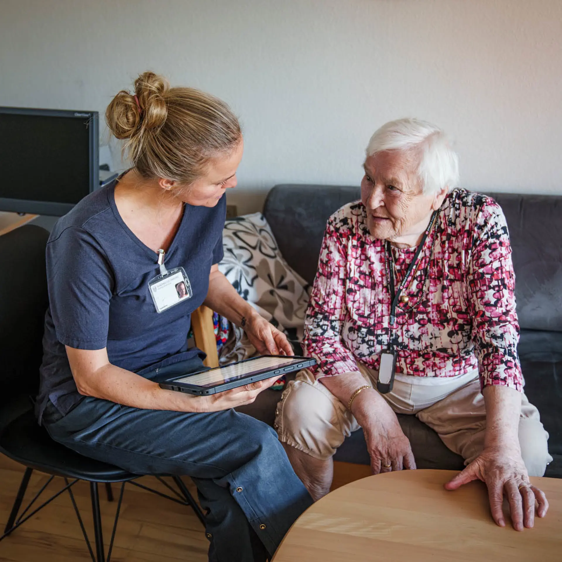 En pleier viser et nettbrett til en eldre kvinne i blomstrete bluse på en svart sofa i en lys stue.