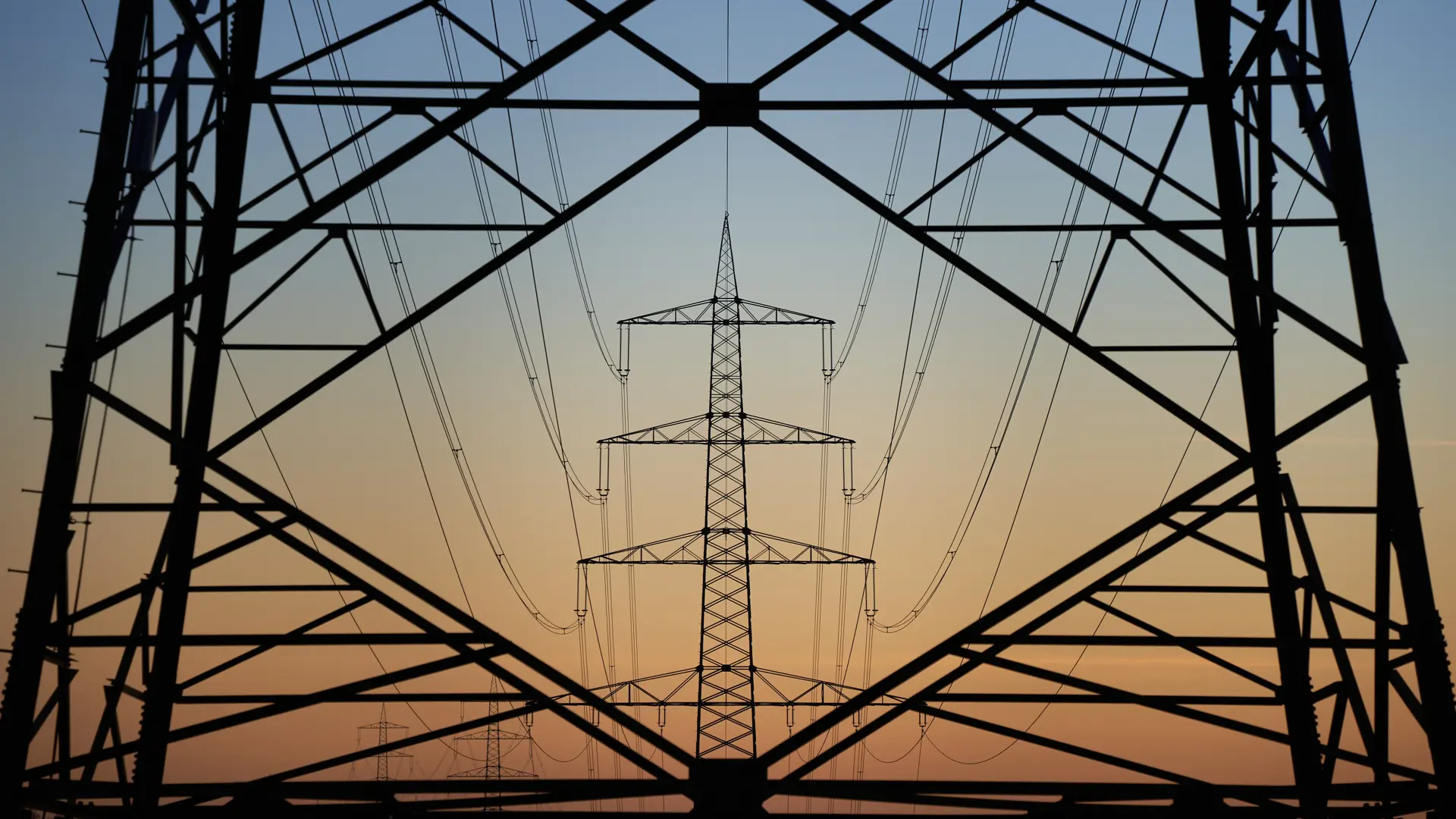Silhouetted high-voltage transmission towers and power lines at sunset, framed by the geometric structure of a foreground pylon.