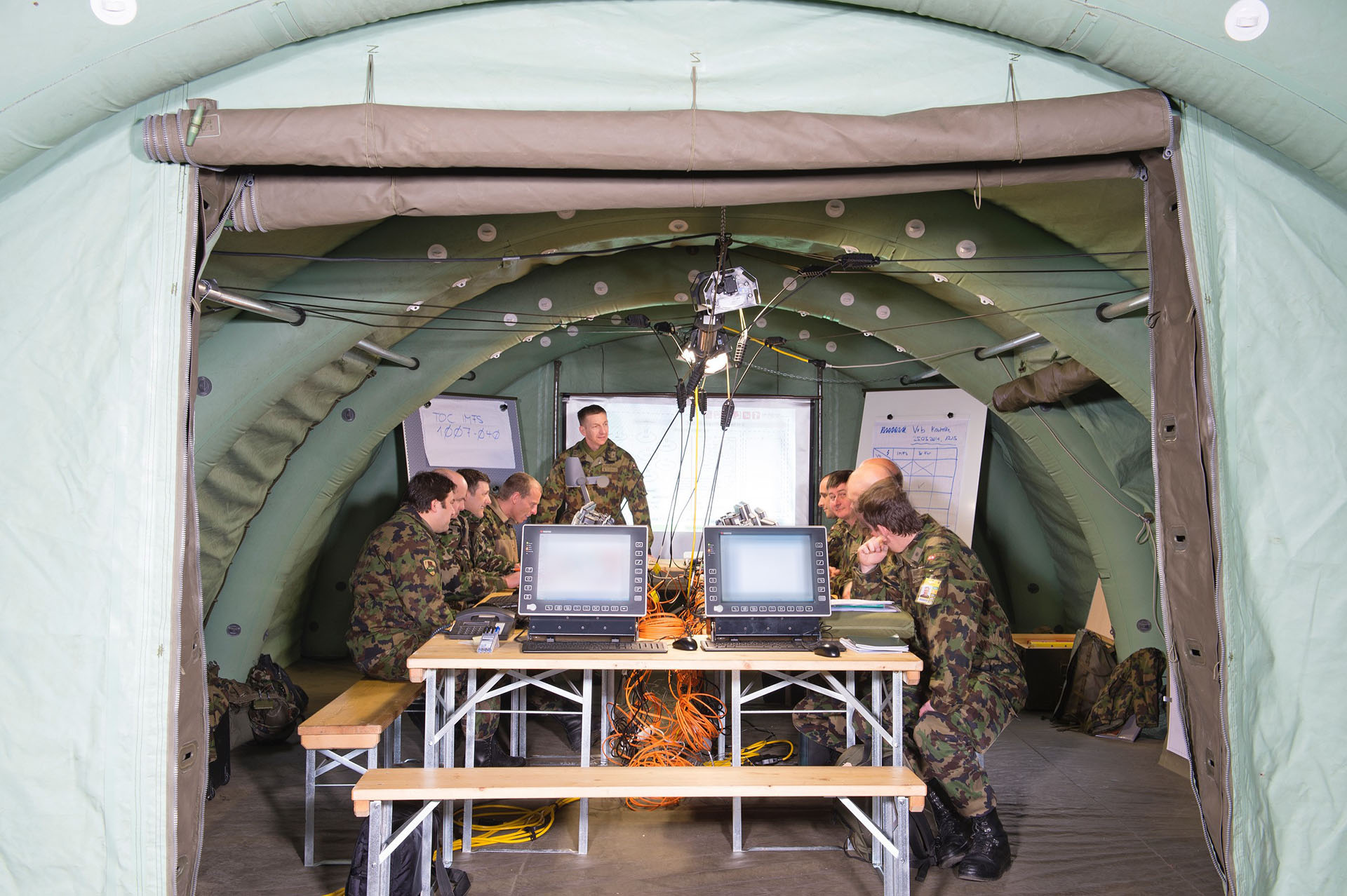 Military personnel in camouflage uniforms conducting a briefing inside a tactical command tent with computers, network cables, and whiteboards.