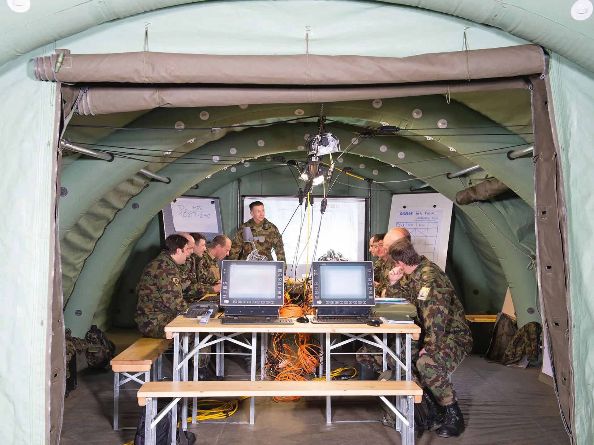 Military personnel in camouflage uniforms conducting a briefing inside a tactical command tent with computers, network cables, and whiteboards.