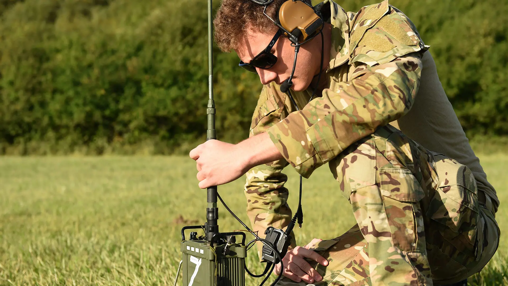 Military electronics technician in camouflage uniform tests high-frequency (HF) radio communications equipment in an outdoor field setting.