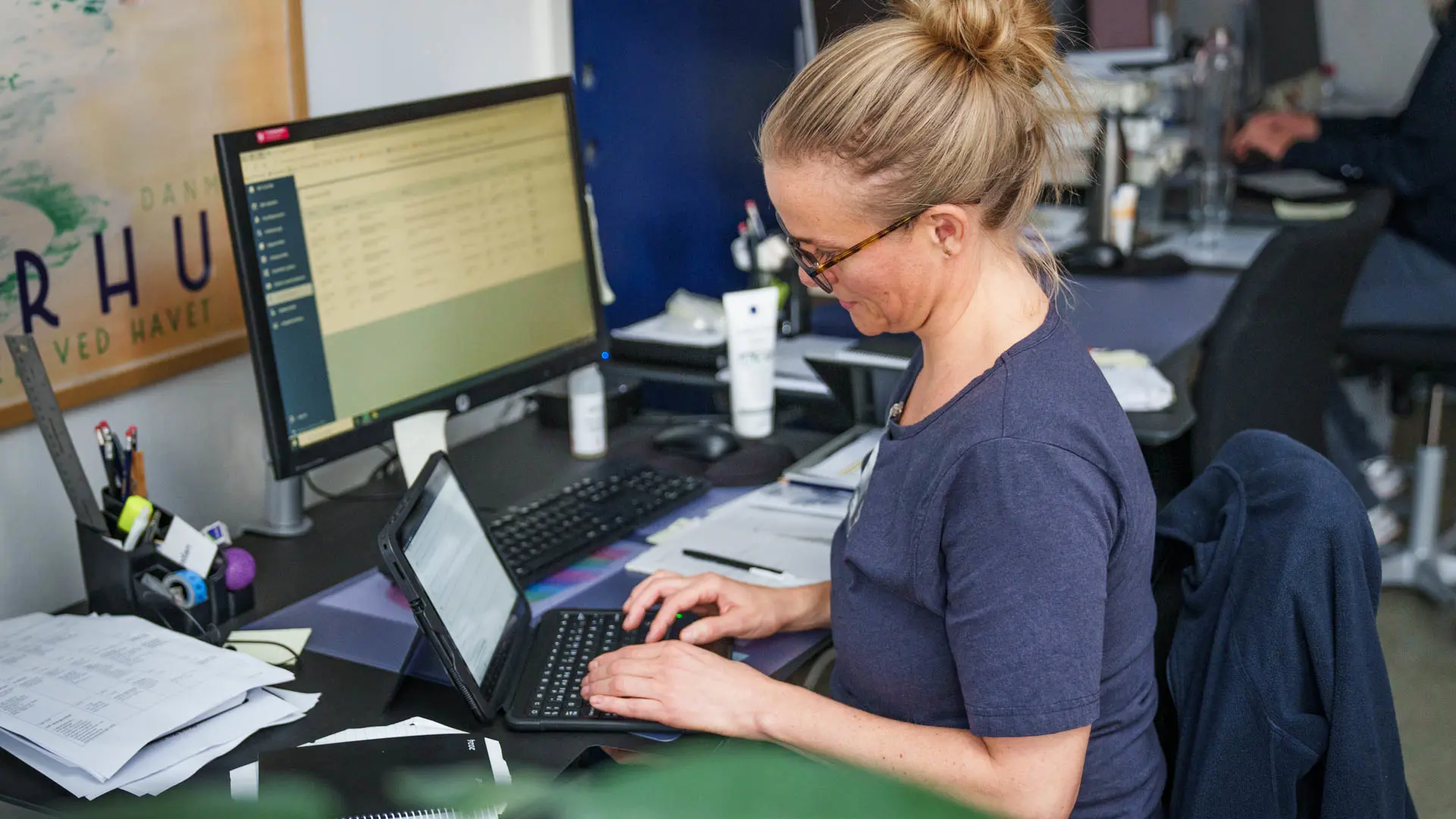 Nurse working at a desk with a tablet and desktop computer, surrounded by documents and medical materials in a clinical office environment.