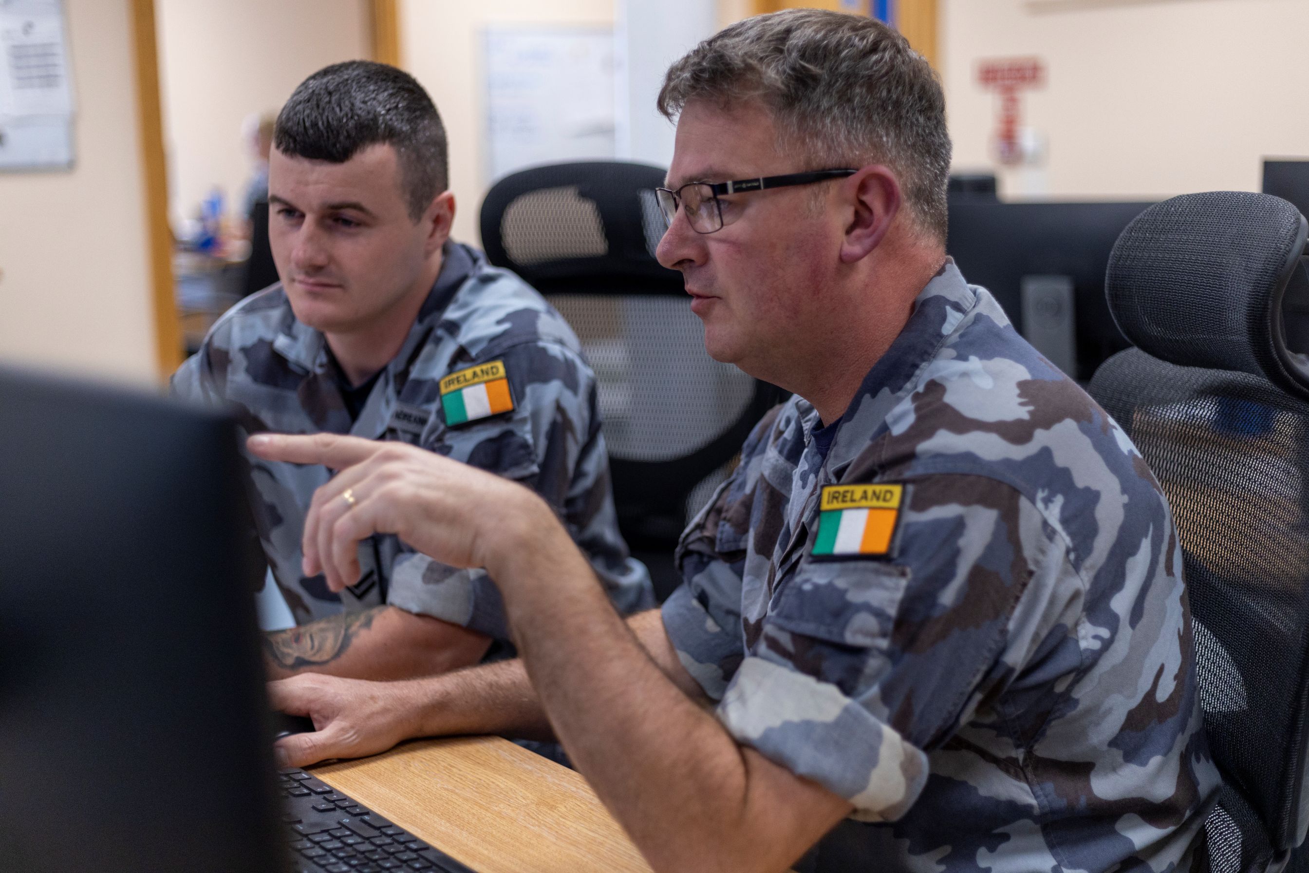 Two Irish military personnel in camouflage uniforms working at a computer workstation, one pointing at the screen.