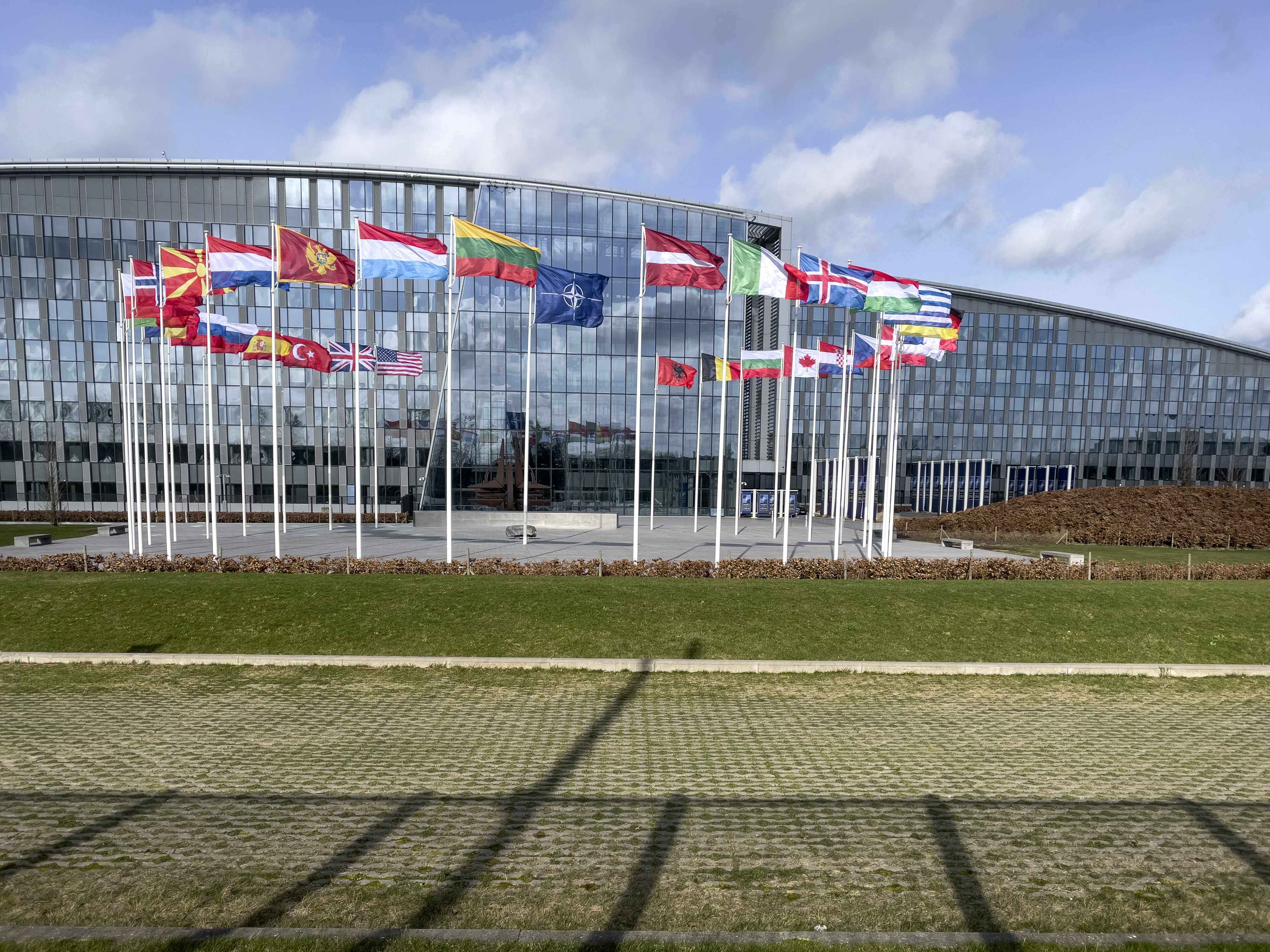 Flags of NATO member countries displayed in front of NATO headquarters in Brussels, Belgium, under a partly cloudy sky.
