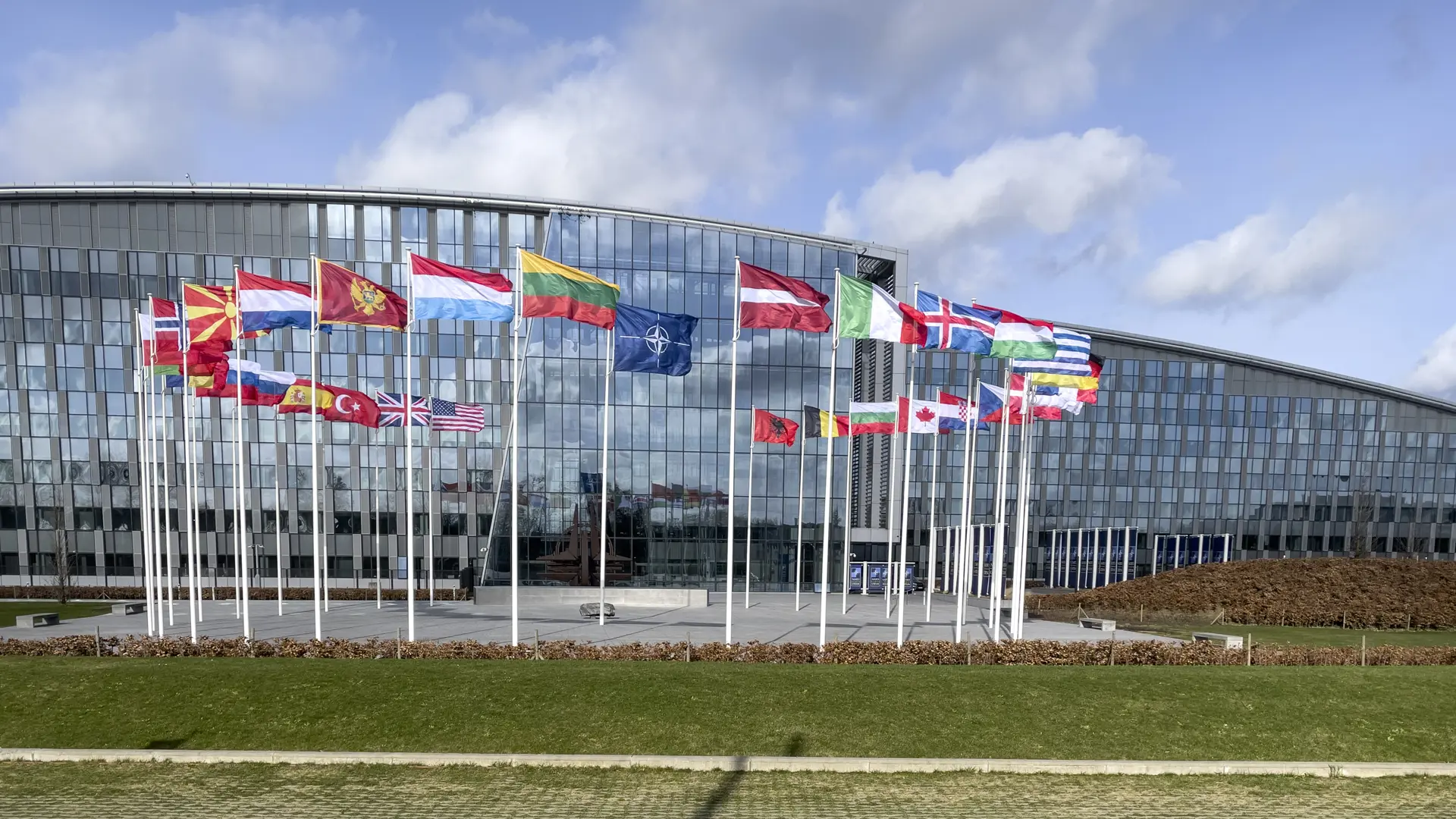 Flags of NATO member countries displayed in front of NATO headquarters in Brussels, Belgium, under a partly cloudy sky.