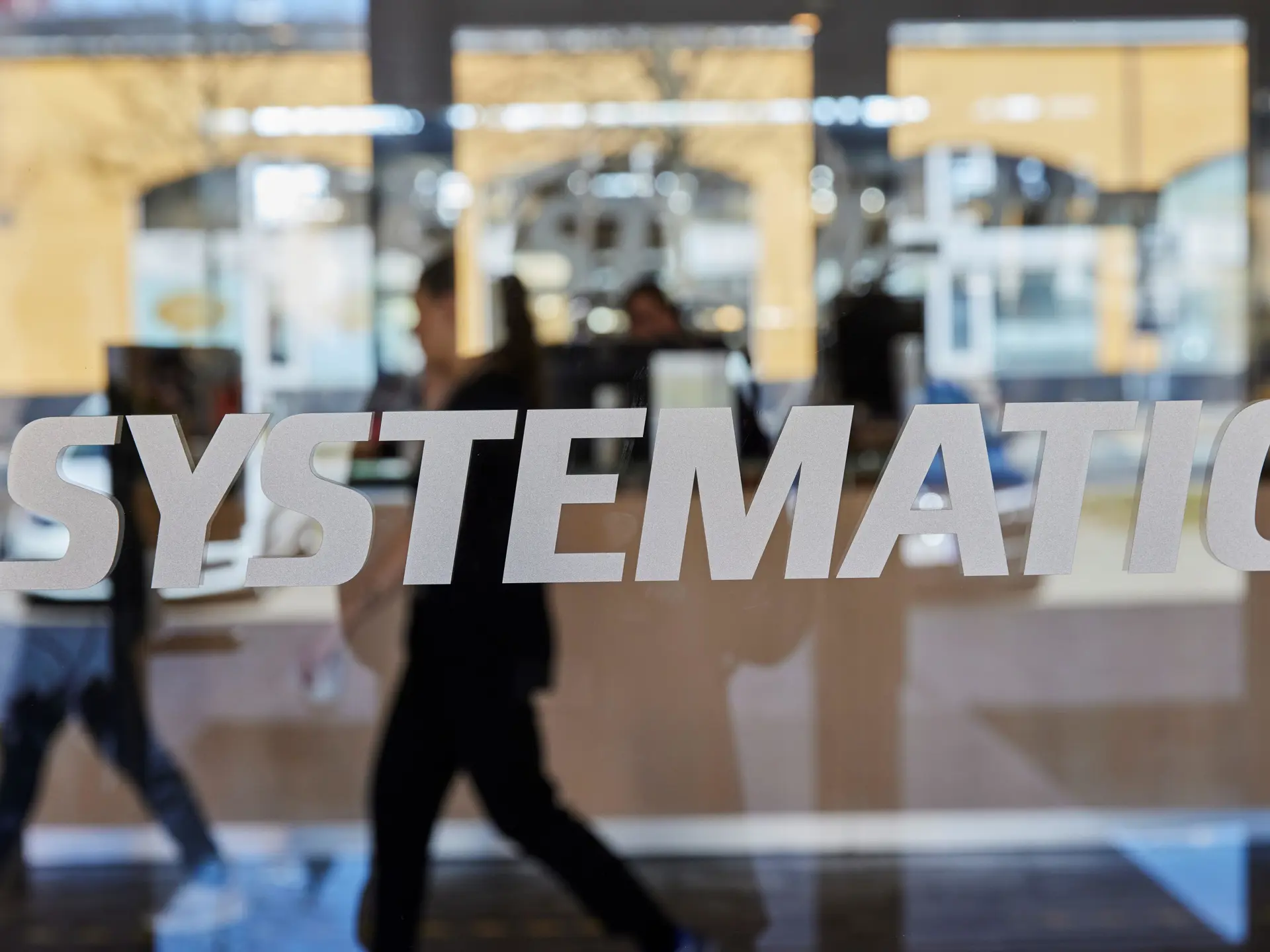 Systematic-logo på en glasvæg i et moderne kontormiljø med medarbejdere, der bevæger sig i baggrunden, som symbol på en professionel software- og teknologivirksomhed.