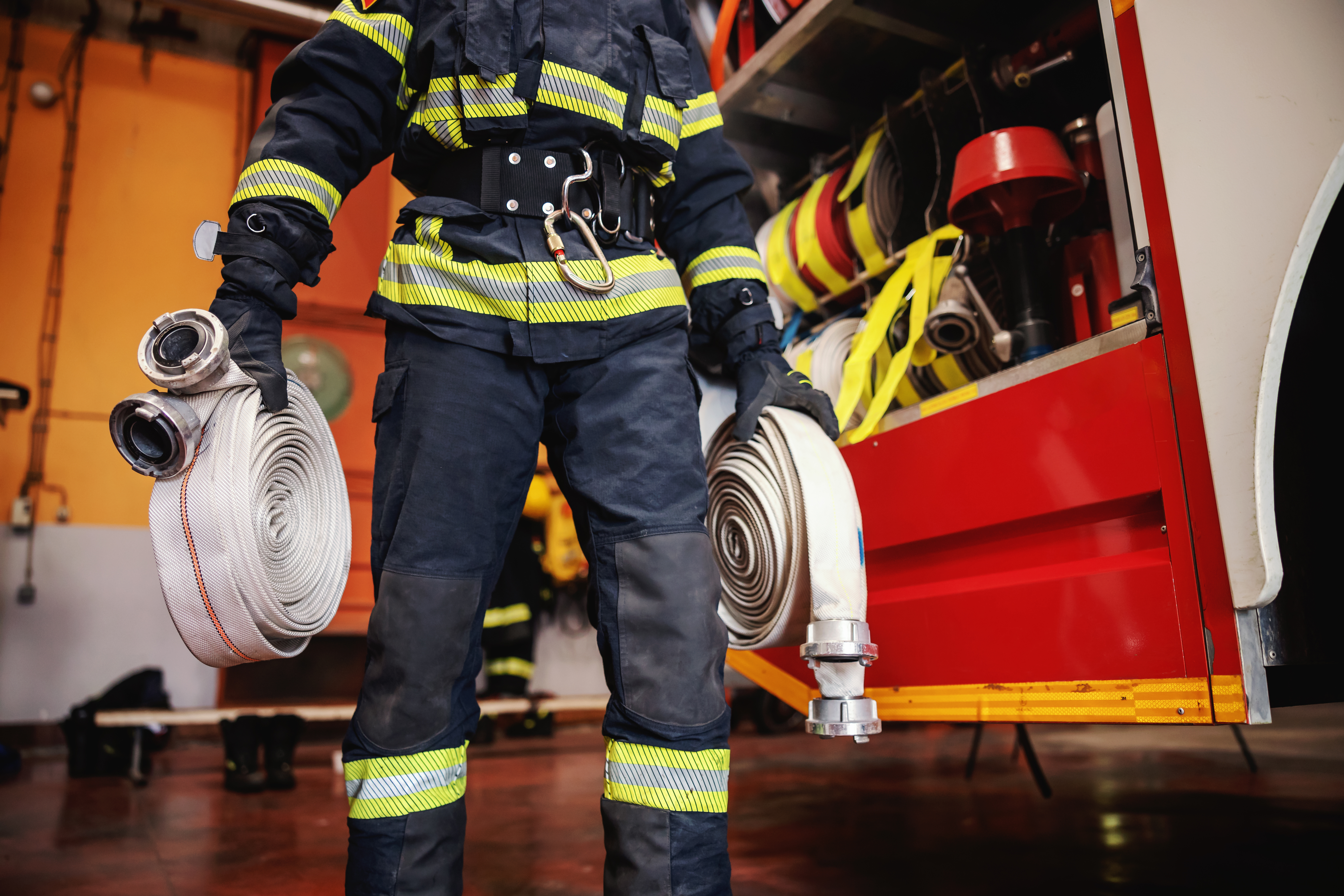 Firefighter in full protective gear holding coiled fire hoses beside a fire truck in the station.