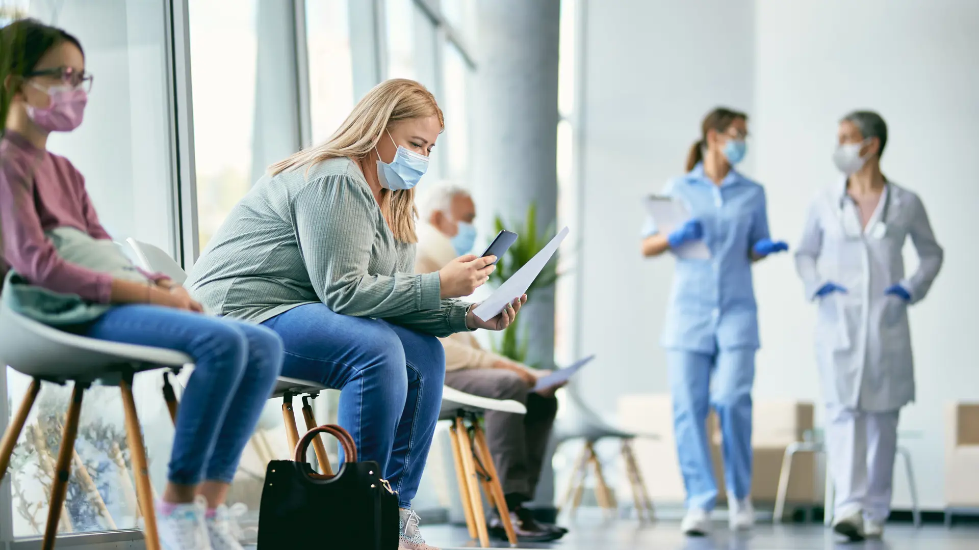 Female patient examining medical paperwork and using mobile phone while sitting in hallway at the hospital