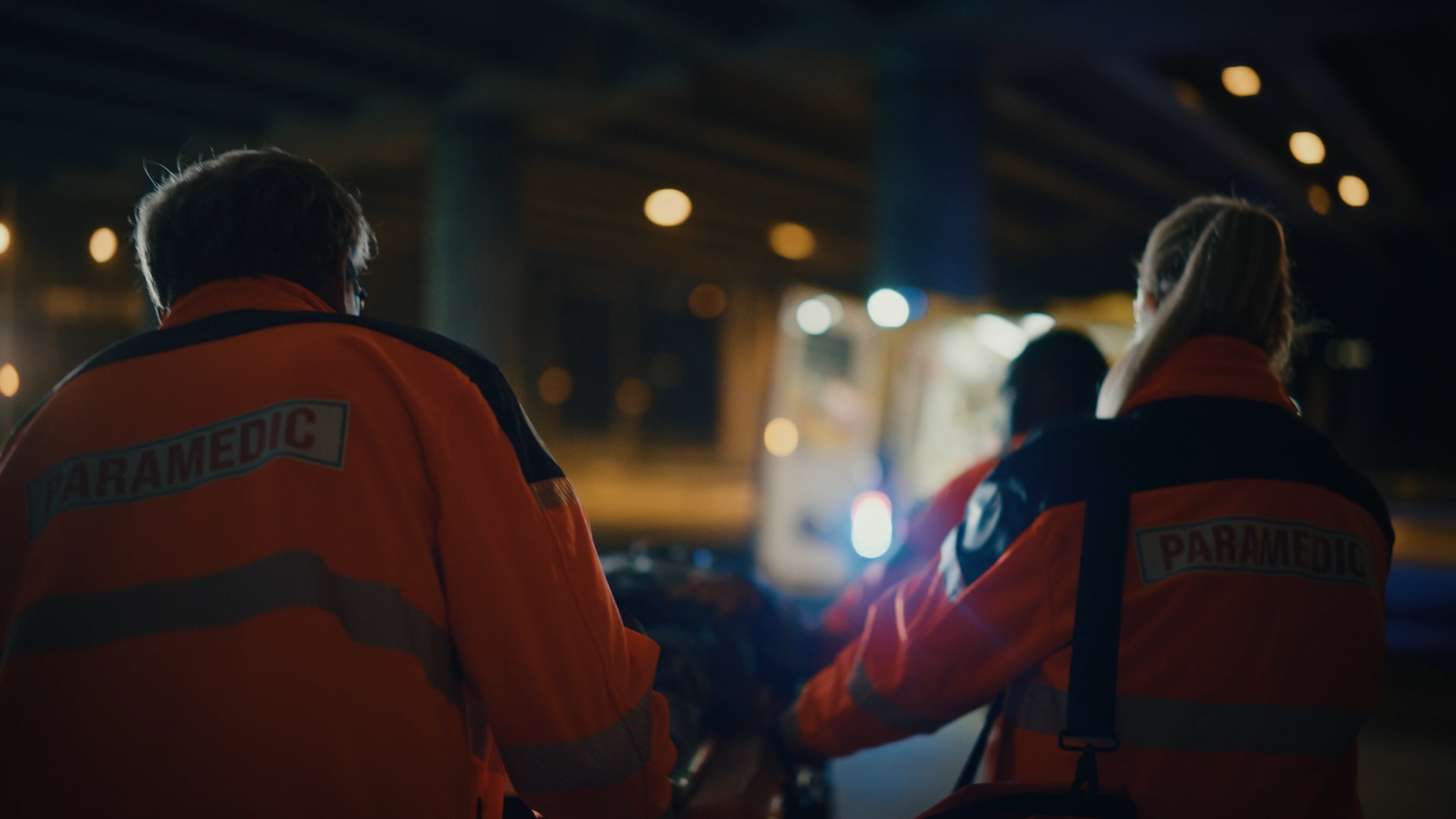 Paramedics in high-visibility jackets transporting a patient on a stretcher toward an ambulance at night.