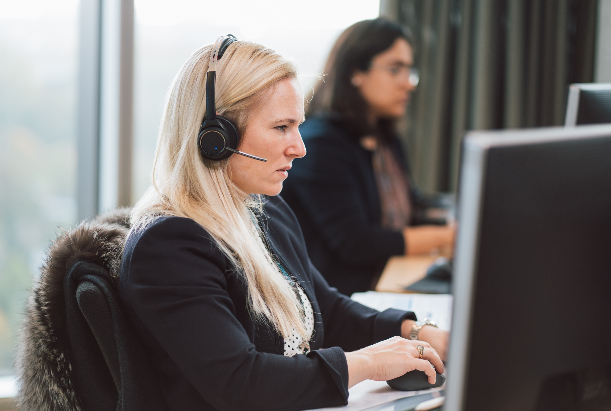 Professional woman wearing a headset, participating in a call while working on a computer in a modern office.