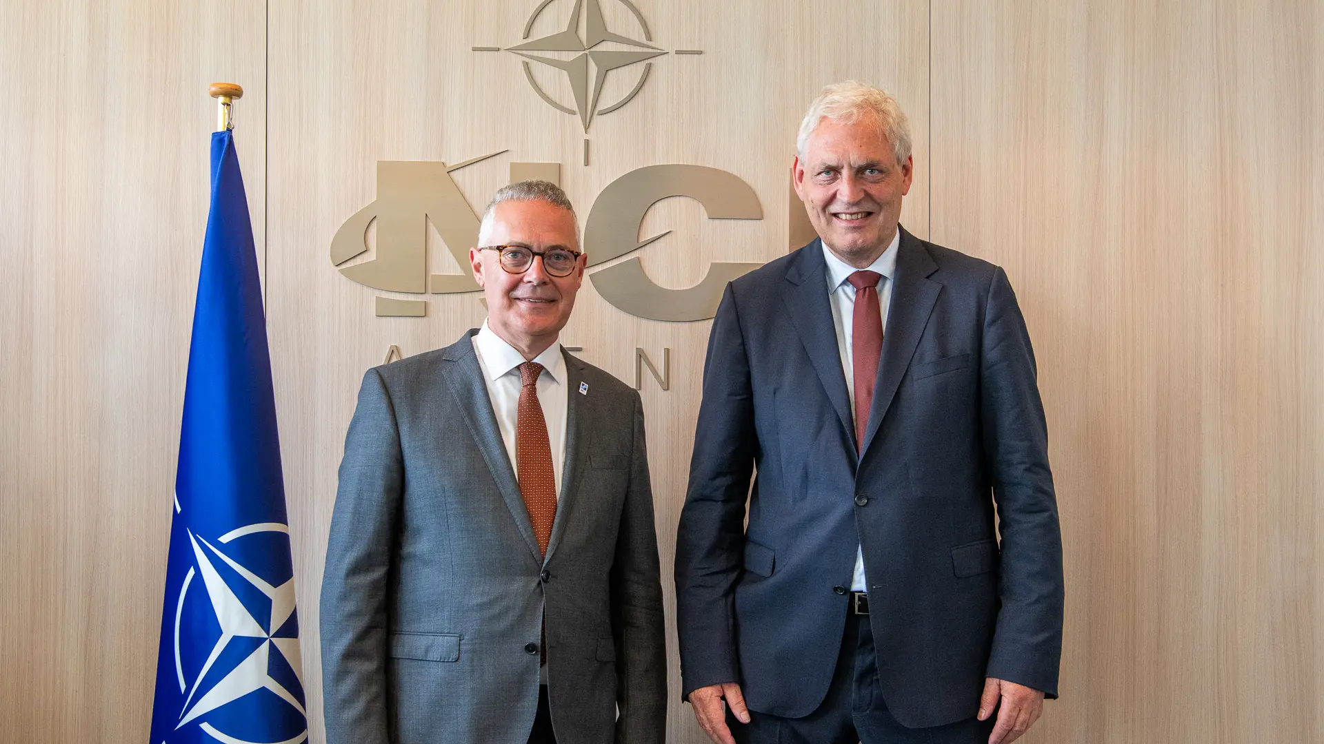 Ludwig Decamps, General Manager of the NCI Agency, and Michael Holm, founder of Systematic, at NATO Headquarters in Brussels, with the NATO flag and NCI Agency logo in the background.