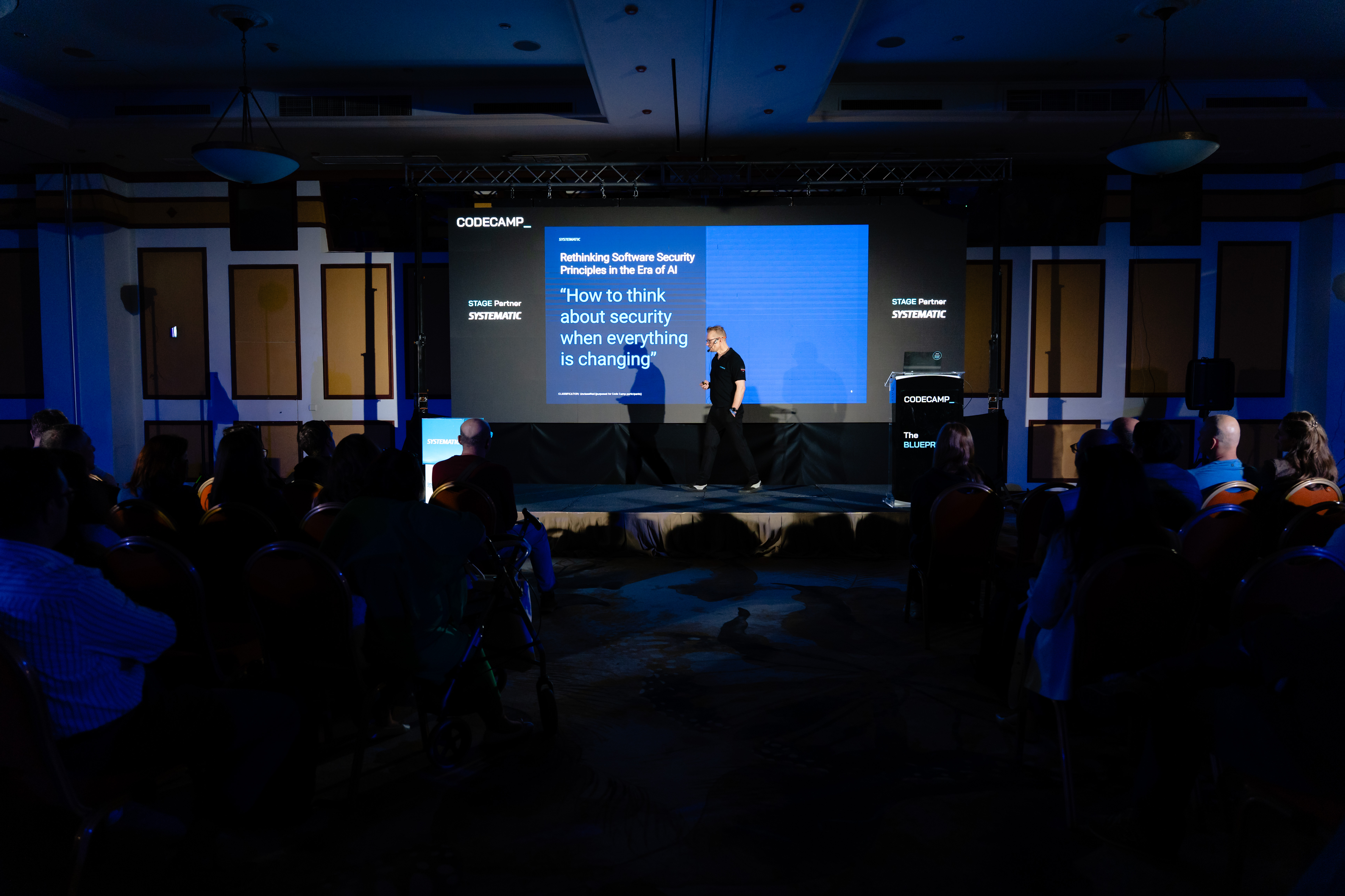 Michael Ustrup speaking on stage at Codecamp, with a Systematic-branded slide on software security in the era of AI projected behind him.