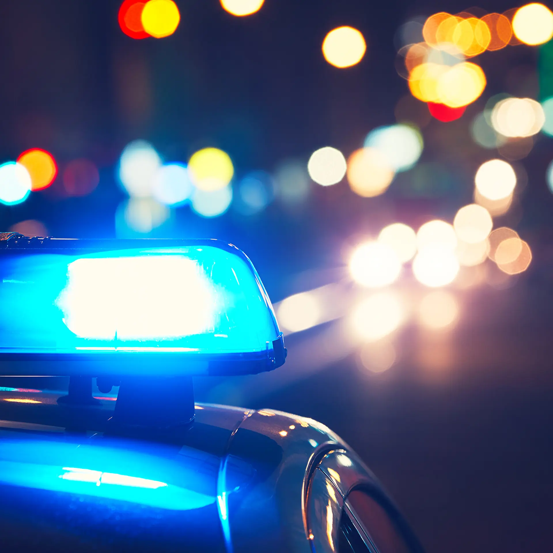 Blue flashing police lights on a patrol car at night with blurred city traffic in the background.