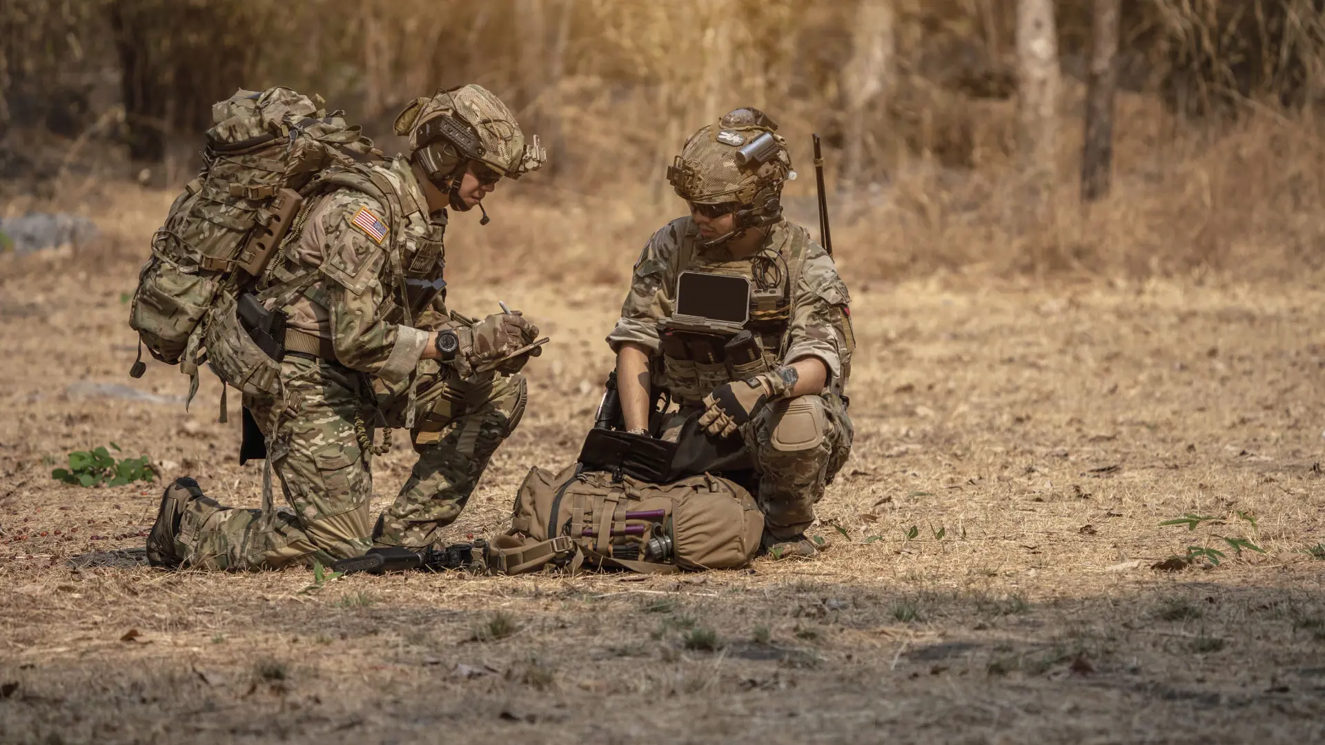 Soldiers in camouflage operate a rugged military device in a dry, wooded area, surrounded by tactical gear.