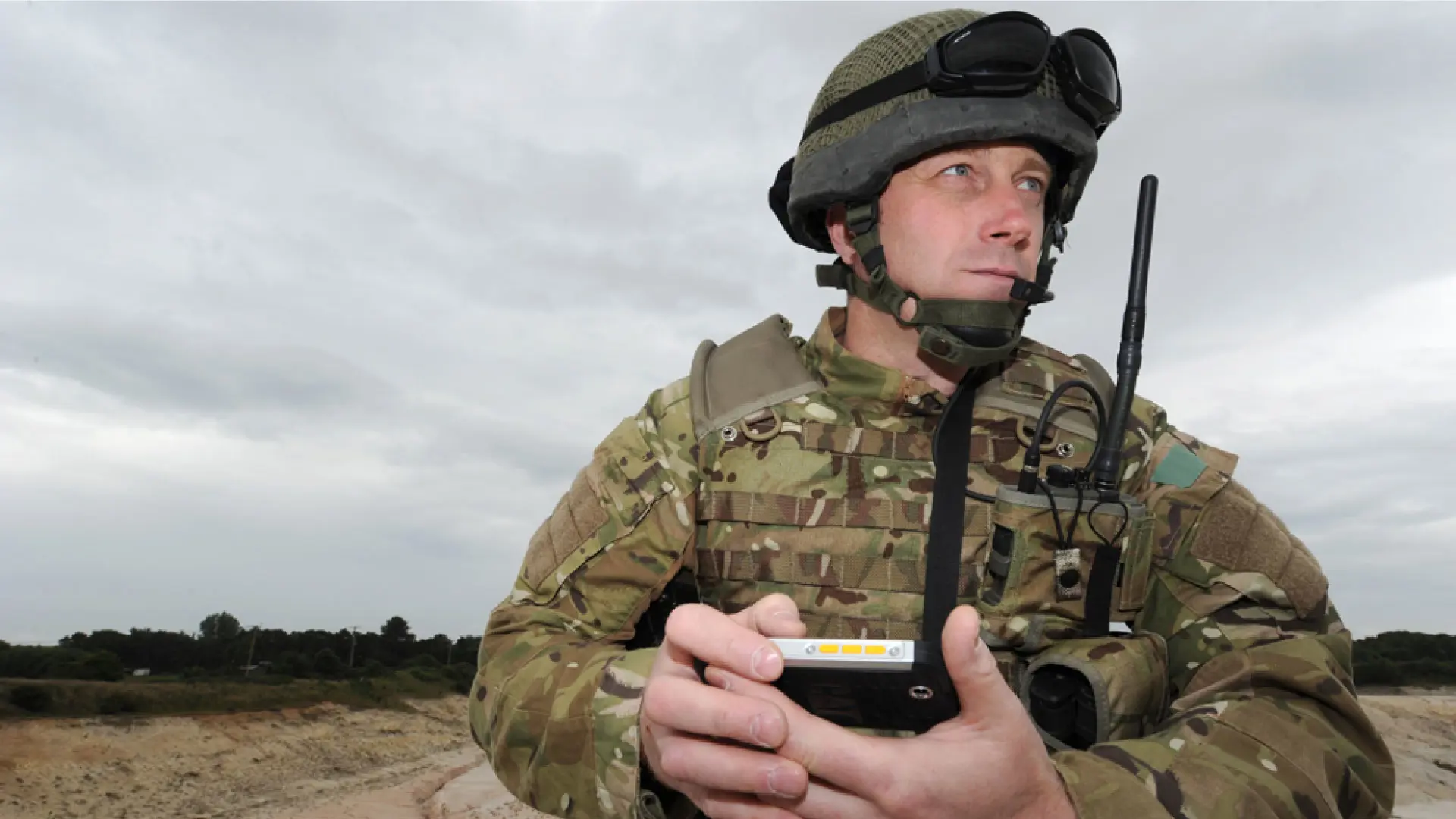 Soldier in camouflage uniform with helmet and radio holds a handheld control device in an open terrain under cloudy skies.
