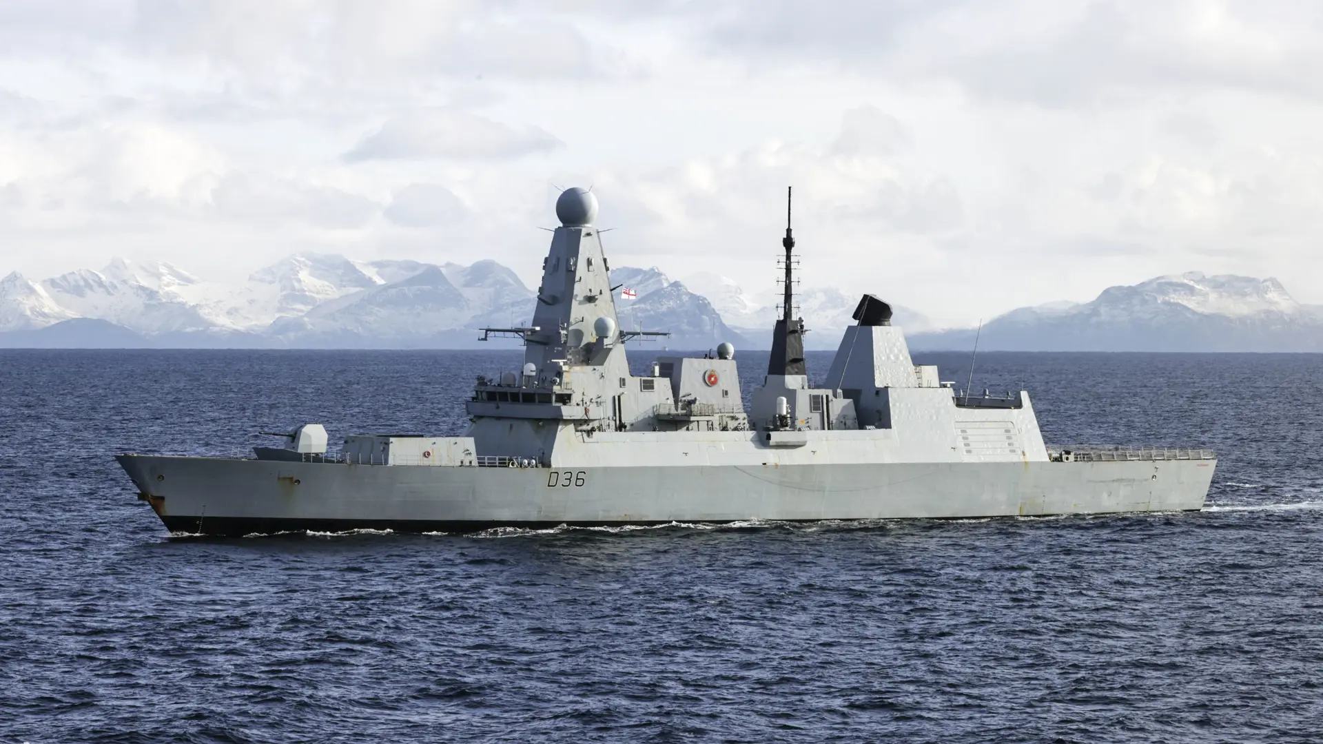 HMS Defender sails through open water with snow-capped mountains in the background