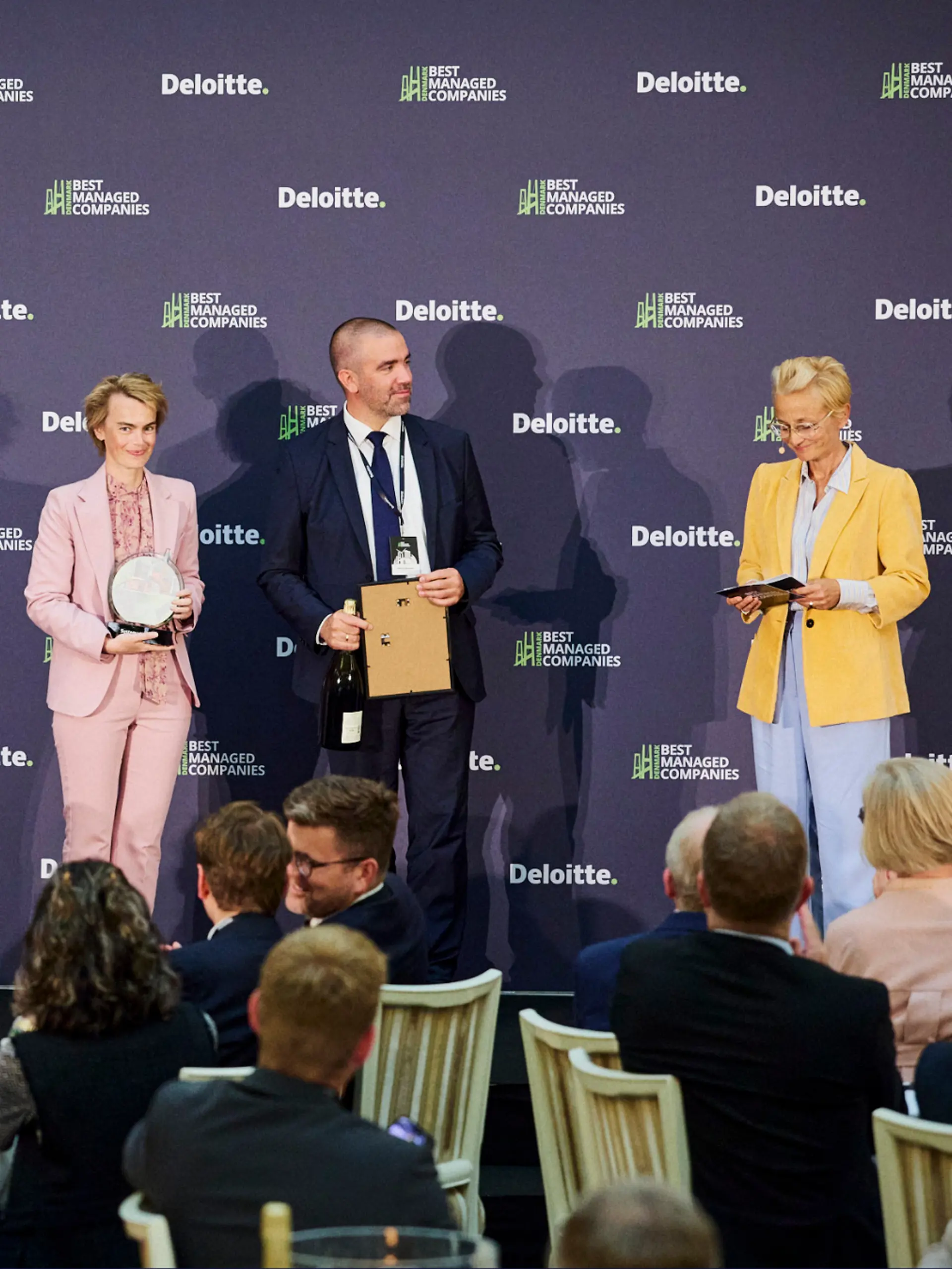 Tre personer står på scenen under Deloittes Best Managed Companies-prisutdeling og holder henholdsvis et trofé, et innrammet diplom og en flaske.