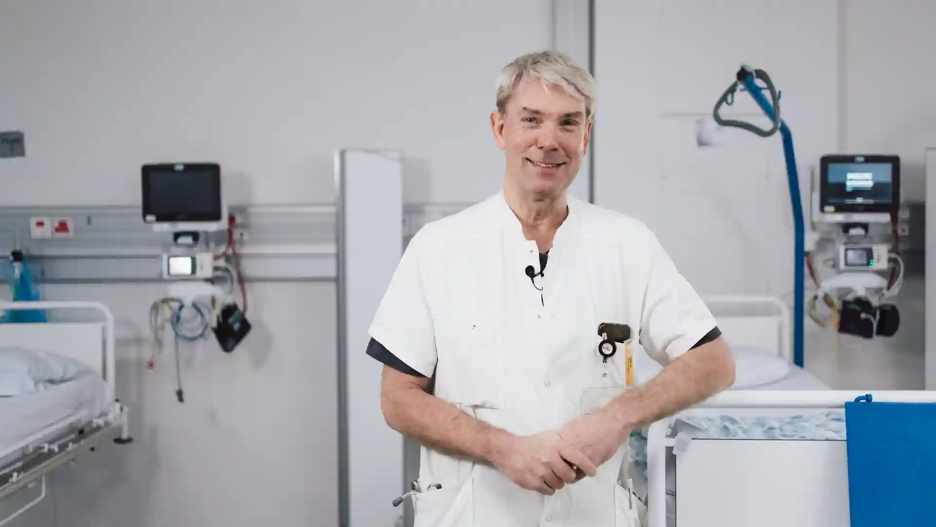 Male healthcare professional standing in a hospital room between two patient beds, smiling at the camera.
