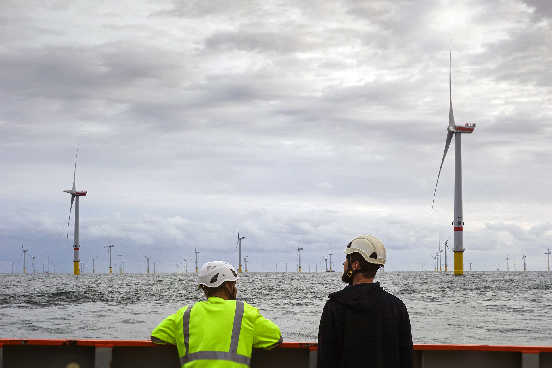 Two offshore wind technicians wearing safety helmets and high-visibility gear observing a large offshore wind farm from a vessel.