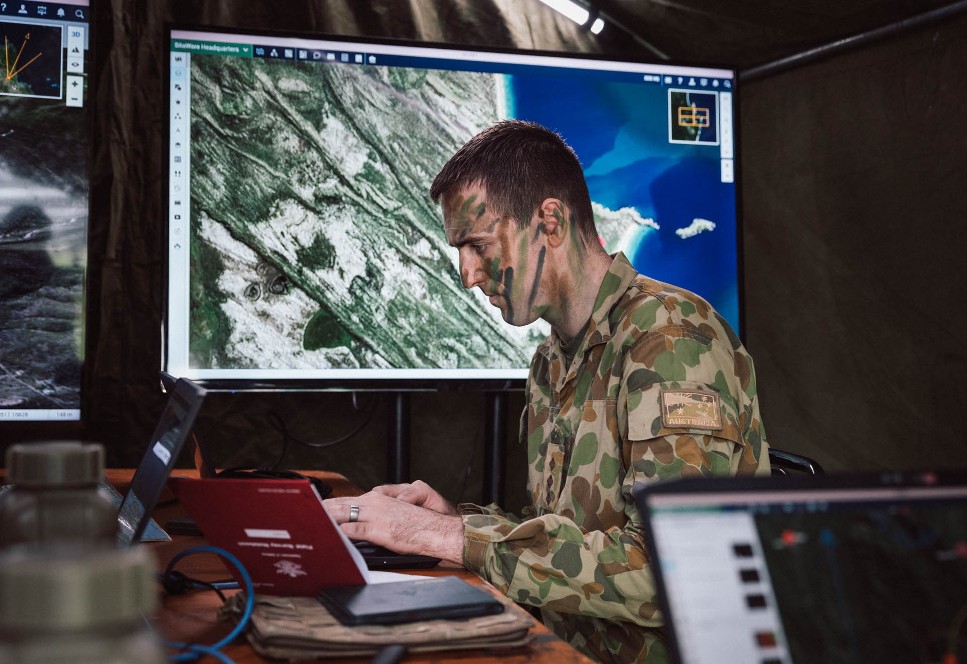 Australian Army soldier in camouflage uniform using a laptop in a field command post, with SitaWare Headquarters displaying a tactical map on screen behind them.