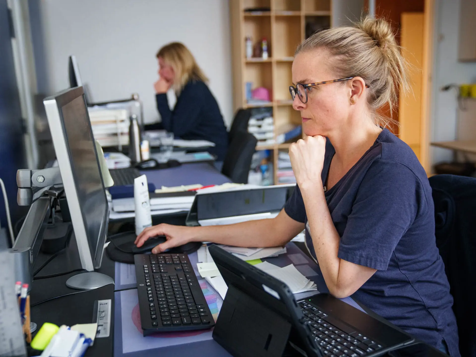 Sygeplejerske arbejder koncentreret ved en stationær computer på et fælleskontor, med tablet og dokumenter på skrivebordet, mens en kollega arbejder i baggrunden.