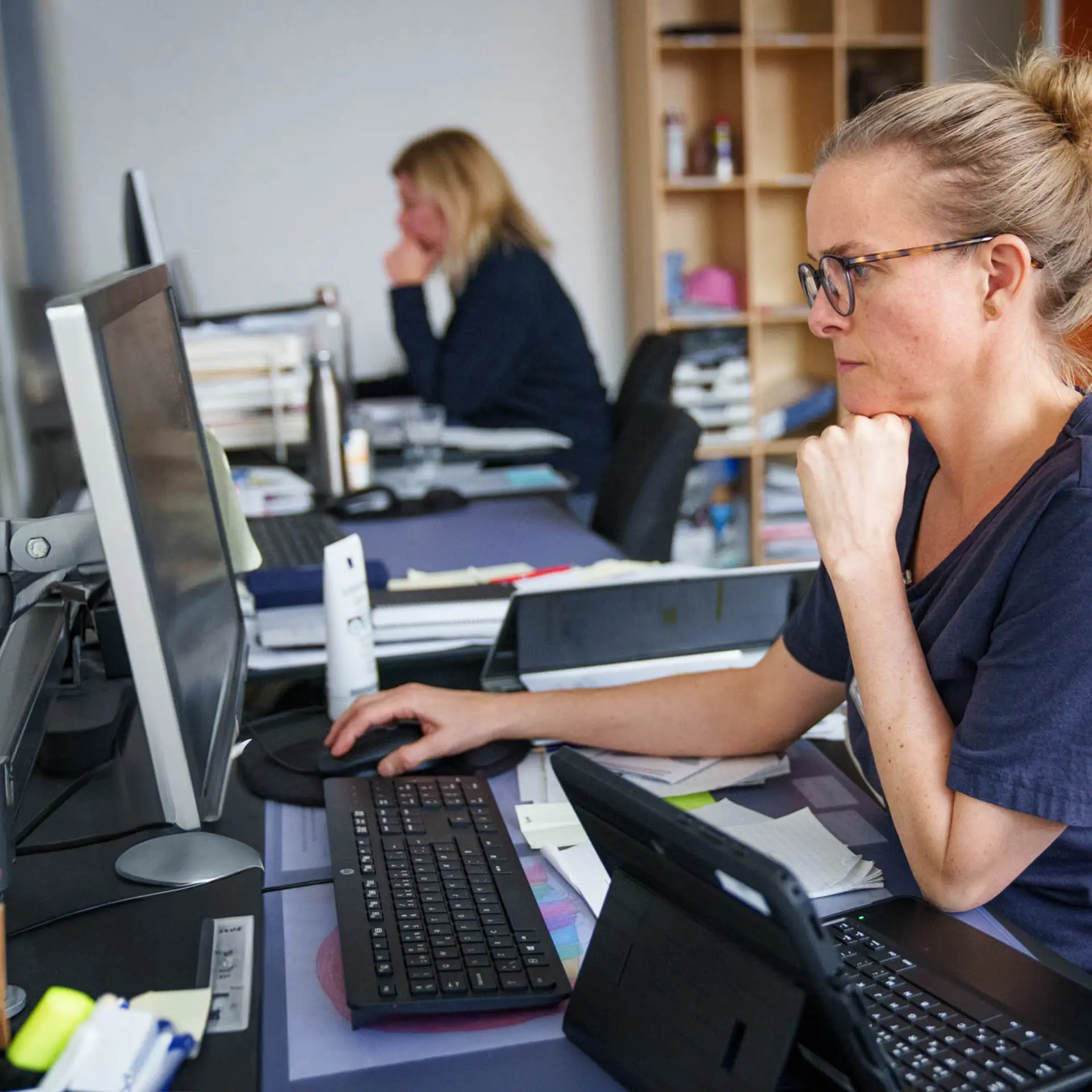 Nurse working intently on a desktop computer at a shared office desk, with a tablet and documents nearby, while a colleague works in the background.