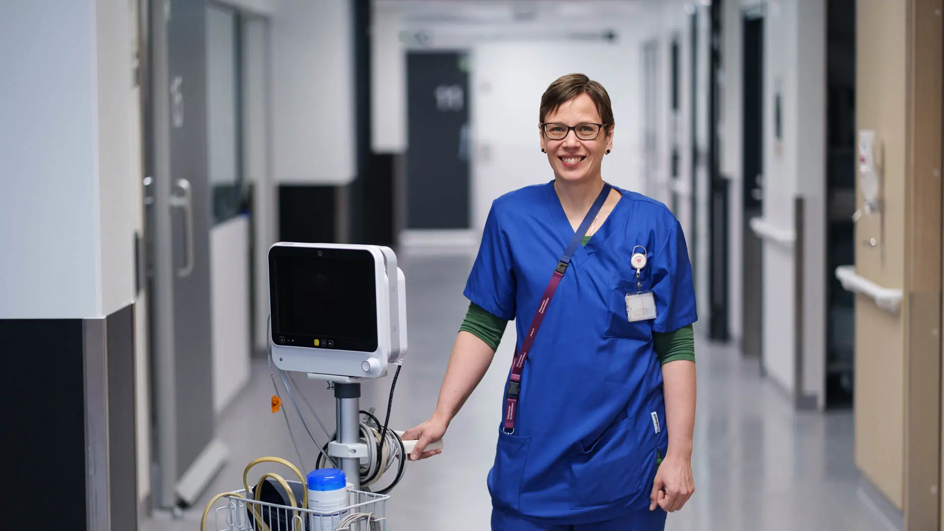 Tiina Syrén, nurse at Tampere University Hospital, standing in a hospital corridor with medical equipment, wearing nursing uniform.