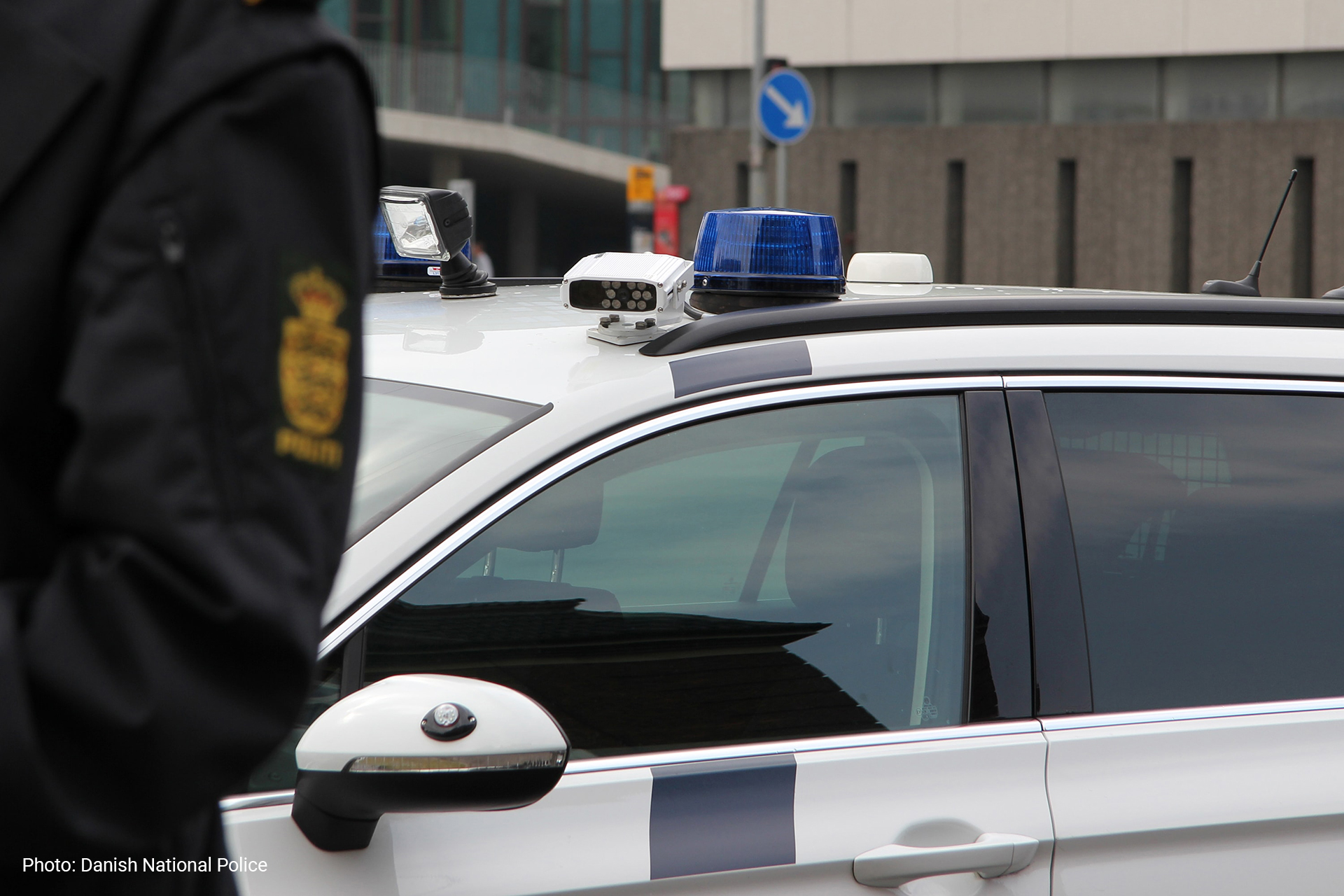 Close-up of a police vehicle with blue lights and roof-mounted equipment, partially obscured by a uniformed police officer standing in the foreground.