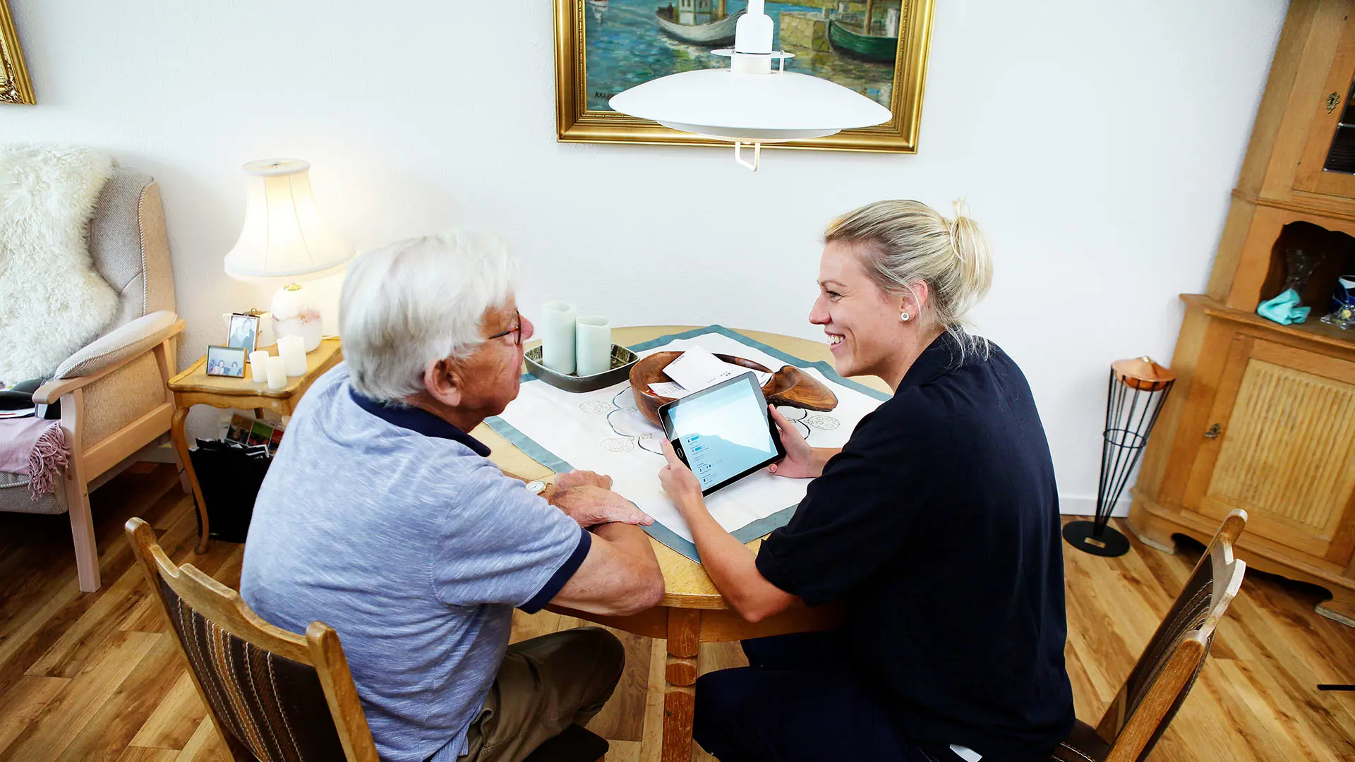 A smiling social worker visits an elderly citizen at home, showing information on a tablet at a dining table in a cozy living room, illustrating digital citizen care.