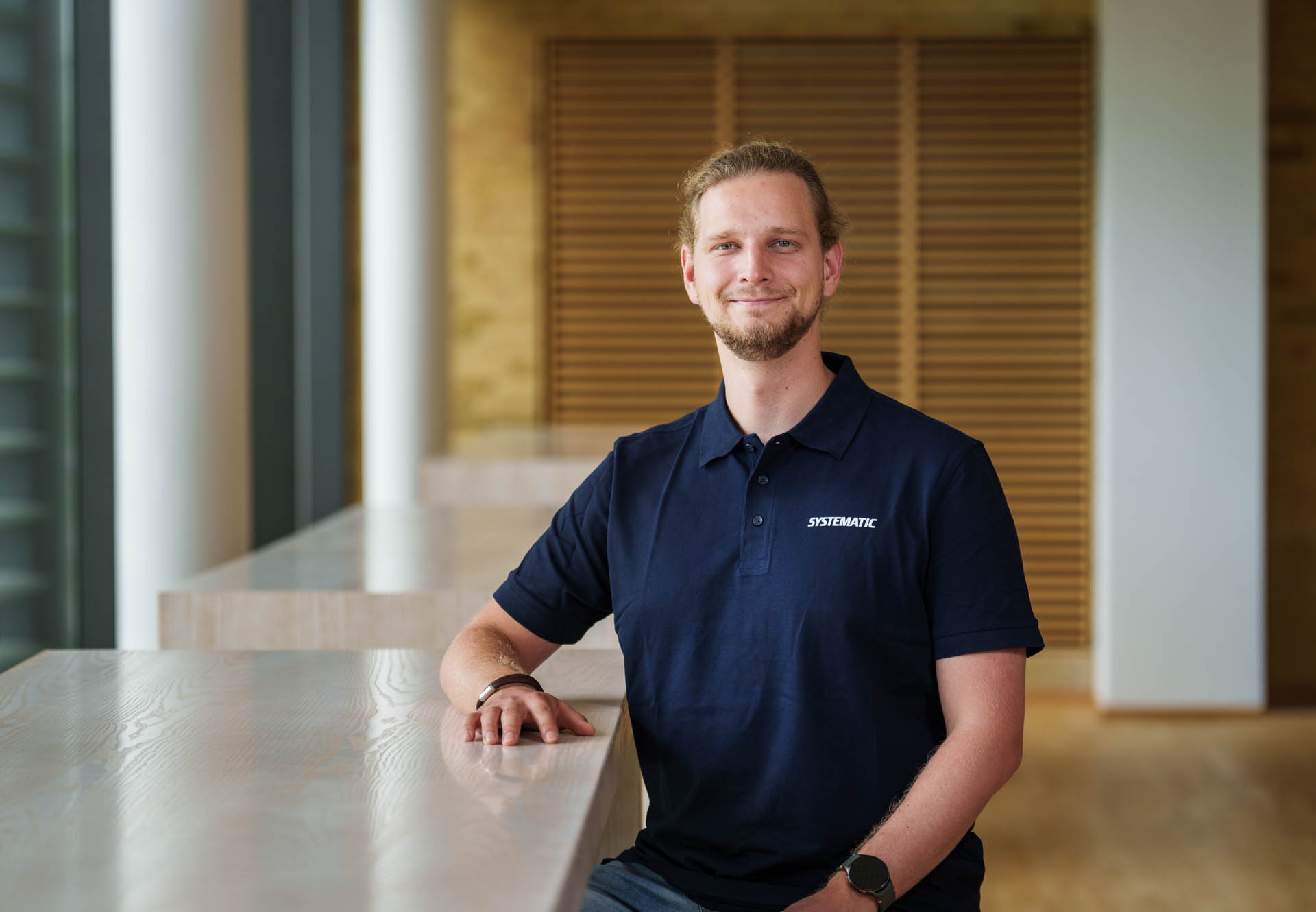 Julian Cornel Berndt wearing a Systematic-branded polo shirt, resting one arm on a wooden counter in an office interior.