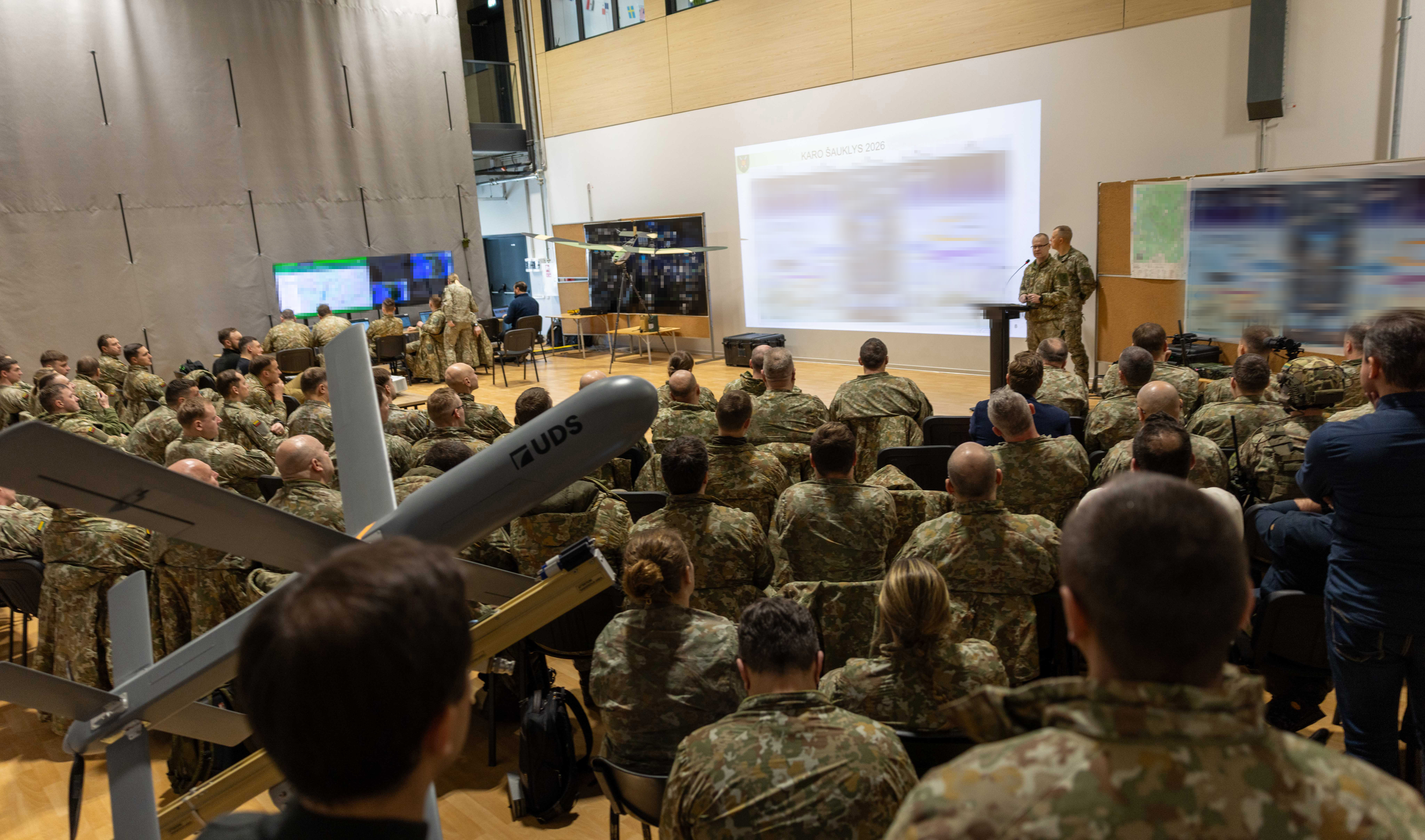 Military personnel in camouflage uniforms attend a briefing at Lithuania's War Herald 2026 exercise, with presenters displaying operational data on a large projection screen in an auditorium setting.