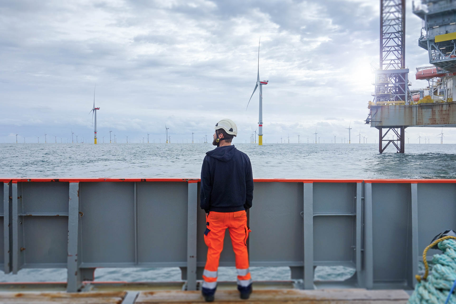 Worker in safety gear standing on a vessel overlooking an offshore wind farm and nearby platform in open sea.