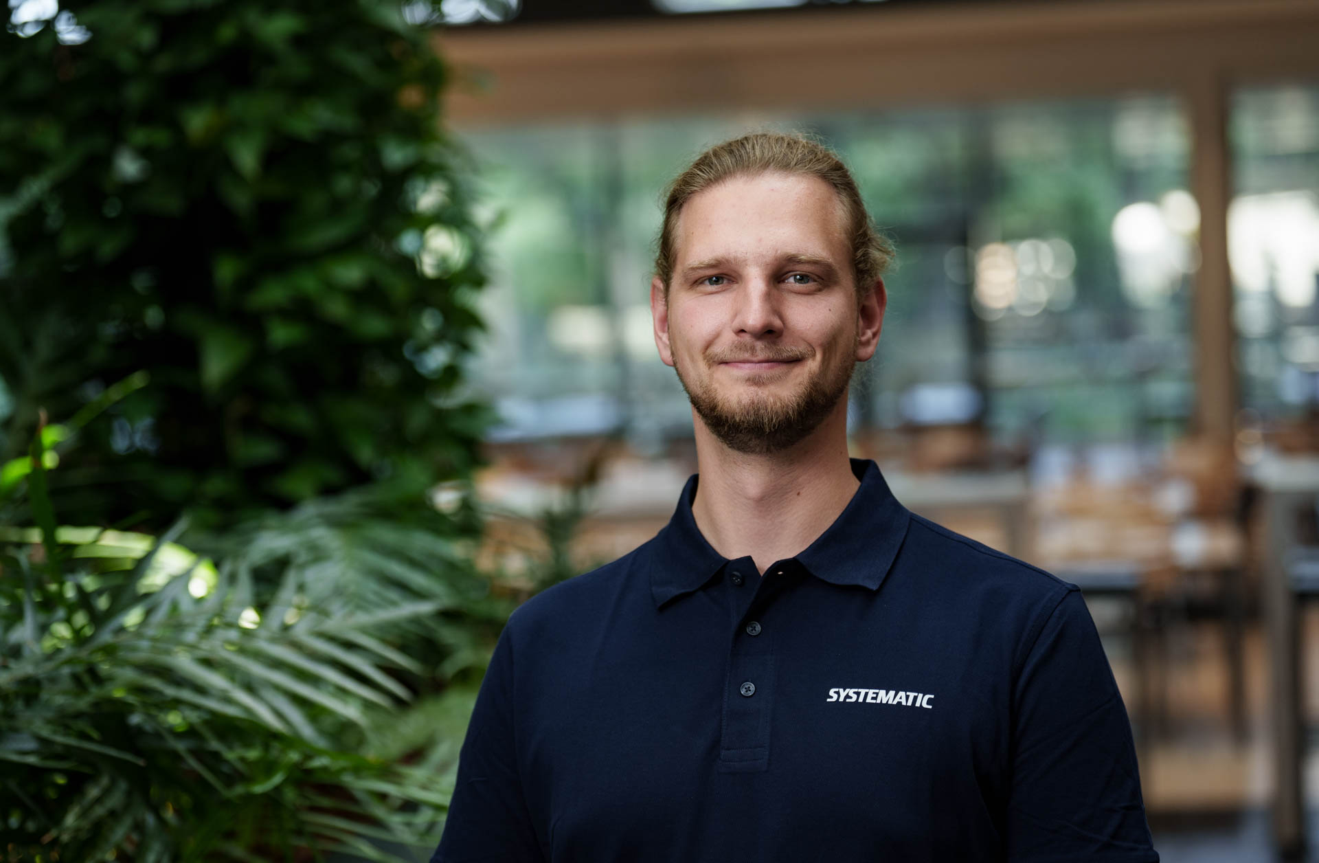 Julian Cornel Berndt wearing a Systematic-branded polo shirt, standing in an office with green plants.