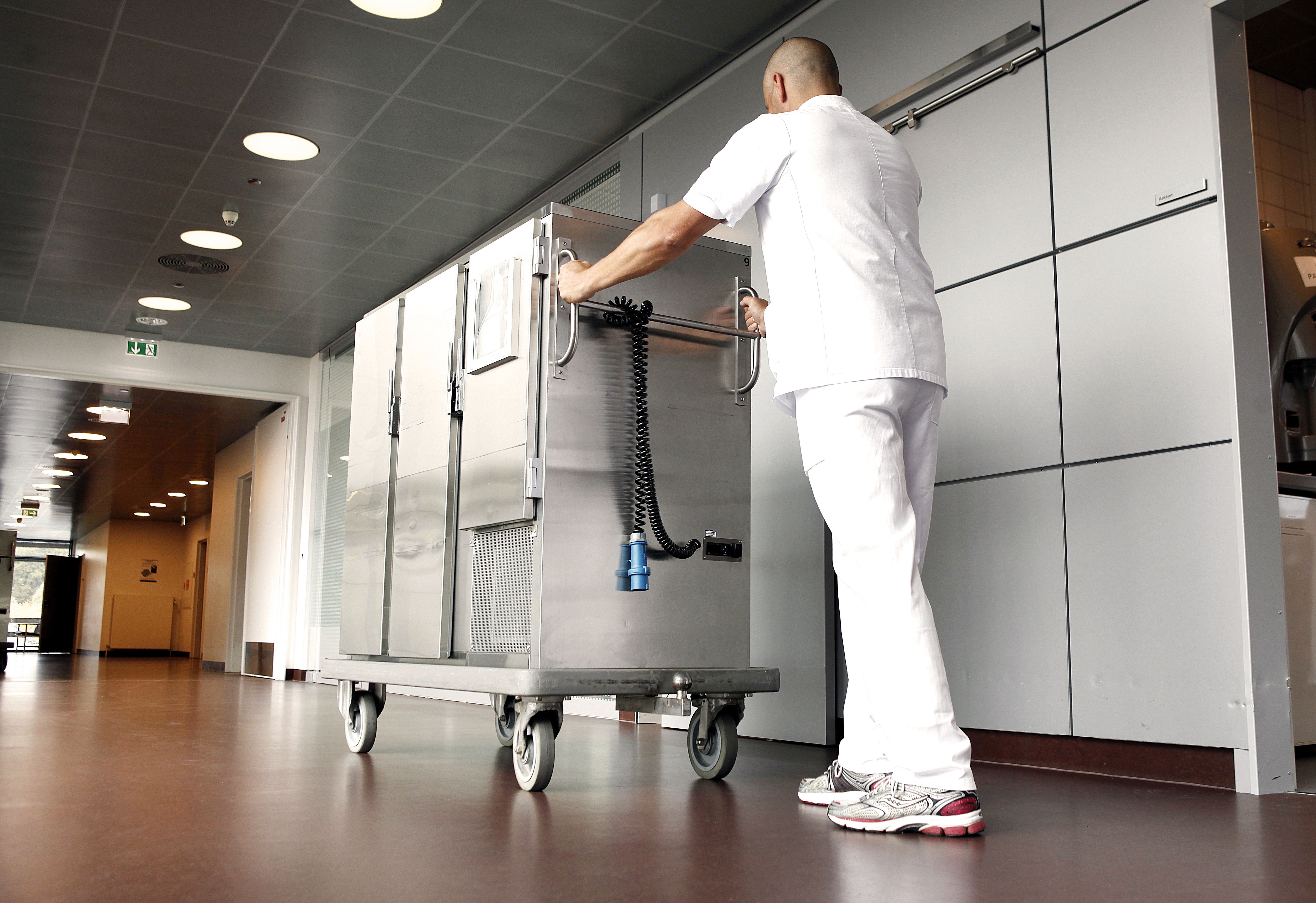 A staff member in a white uniform pushes a large stainless steel trolley along a corridor.