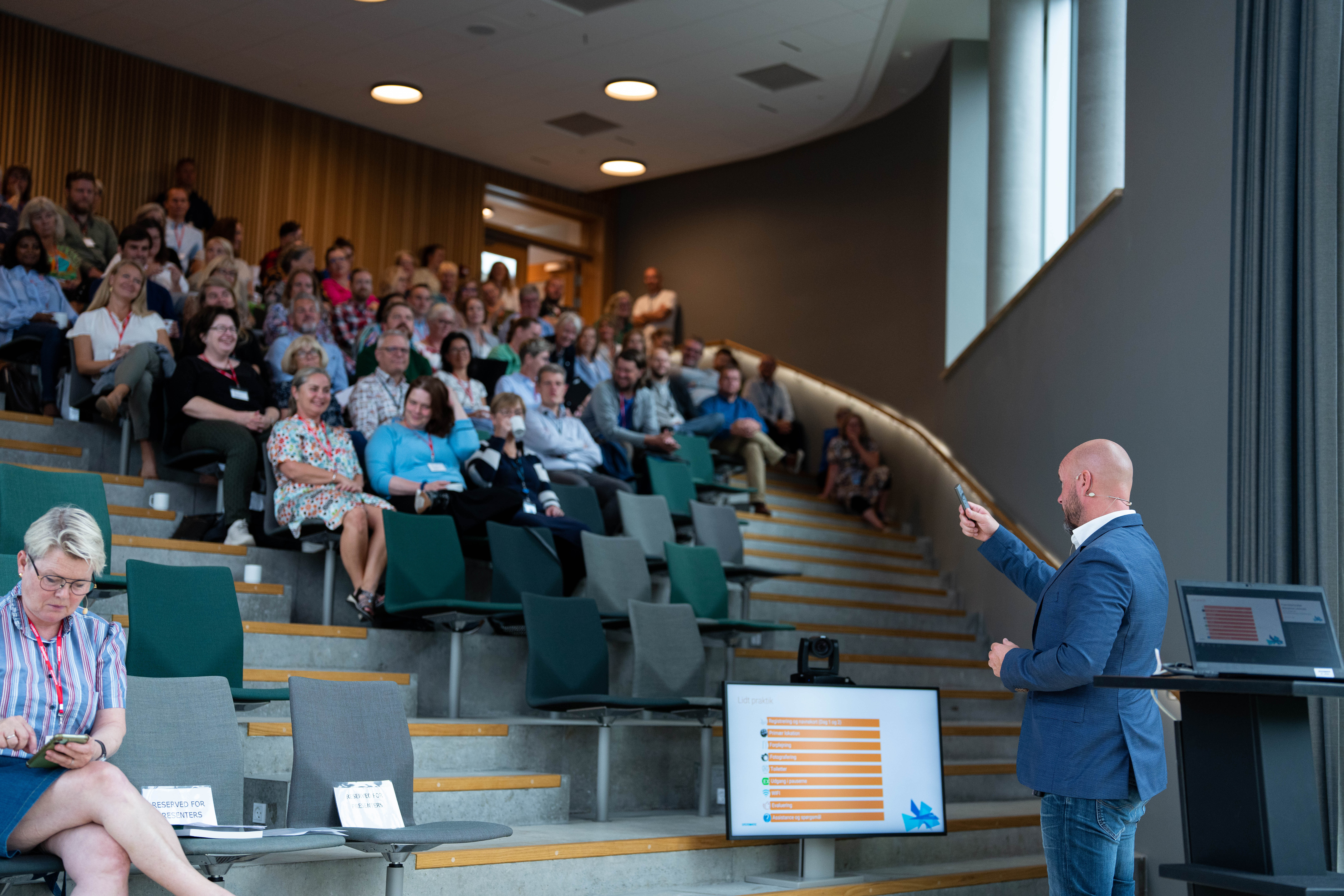 En oplægsholder præsenterer for et publikum i et moderne auditorium, hvor deltagerne sidder på trappetrin og følger med i præsentationen vist på en skærm.