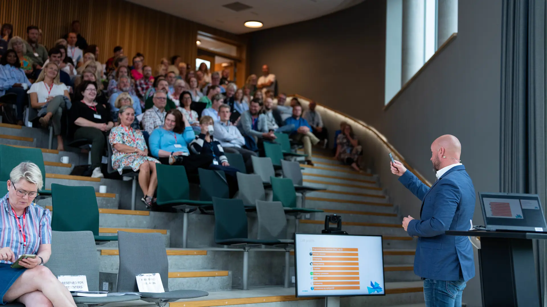 En oplægsholder præsenterer for et publikum i et moderne auditorium, hvor deltagerne sidder på trappetrin og følger med i præsentationen vist på en skærm.