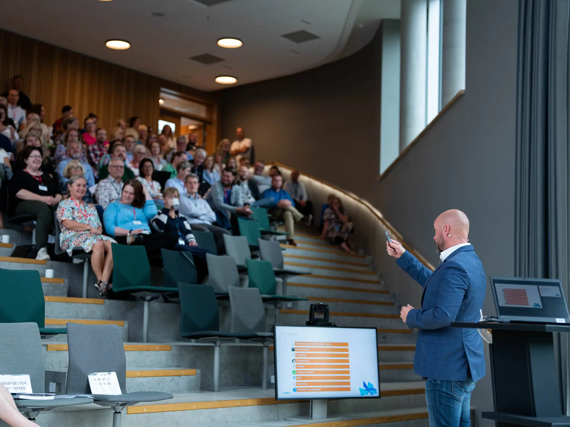 En oplægsholder præsenterer for et publikum i et moderne auditorium, hvor deltagerne sidder på trappetrin og følger med i præsentationen vist på en skærm.