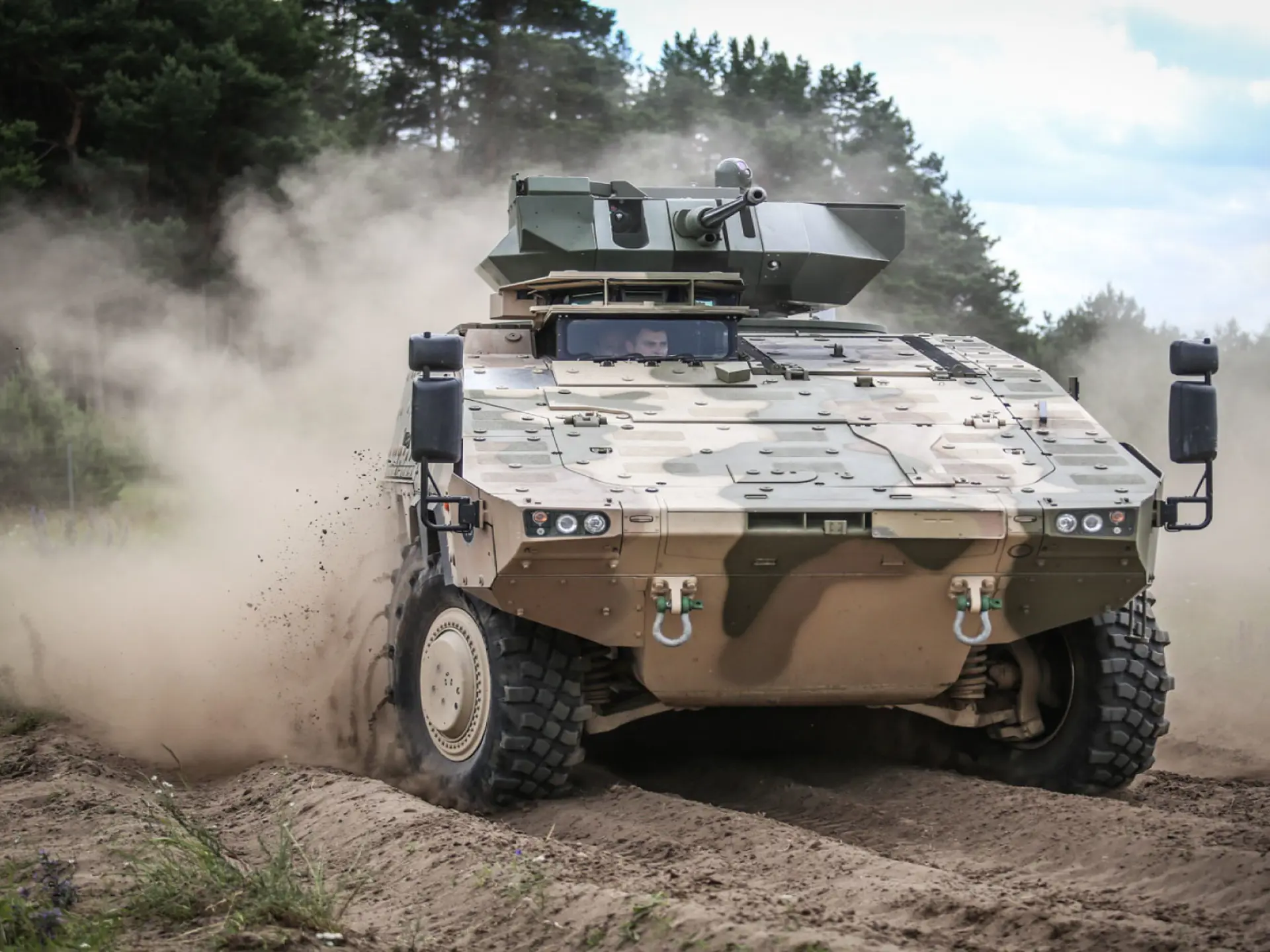 Armored military vehicle kicks up dust while driving off-road through a forested training area.v