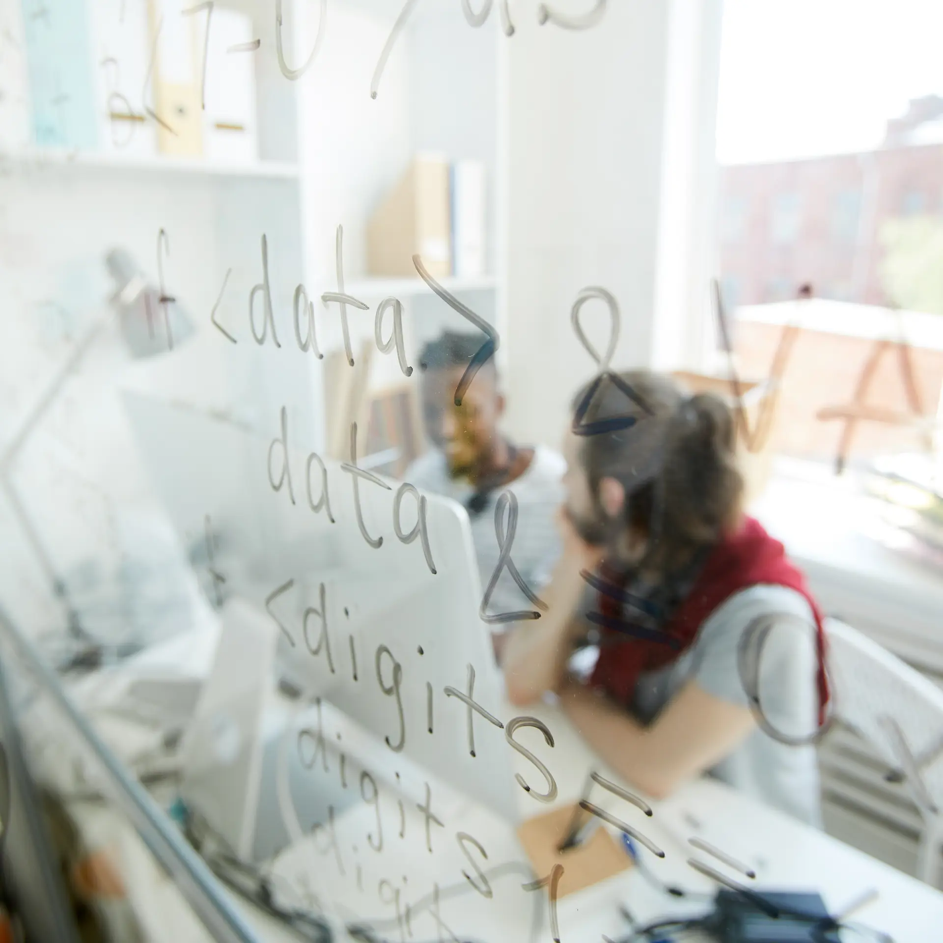 Two developers working behind a glass wall covered in programming syntax and code snippets, in a bright, modern office setting.