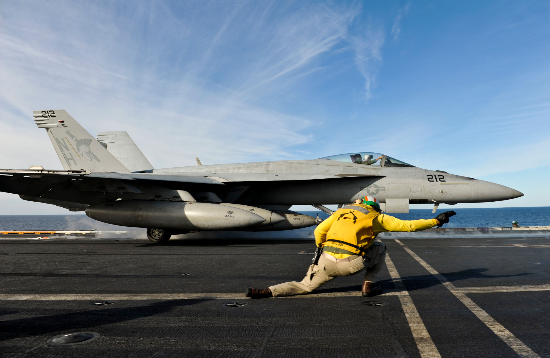 Ein Besatzungsmitglied auf dem Flugdeck eines Flugzeugträgers signalisiert einem F/A-18-Hornet-Kampfjet beim Startvorbereitungen – ein Beispiel für Marineflugbetrieb.