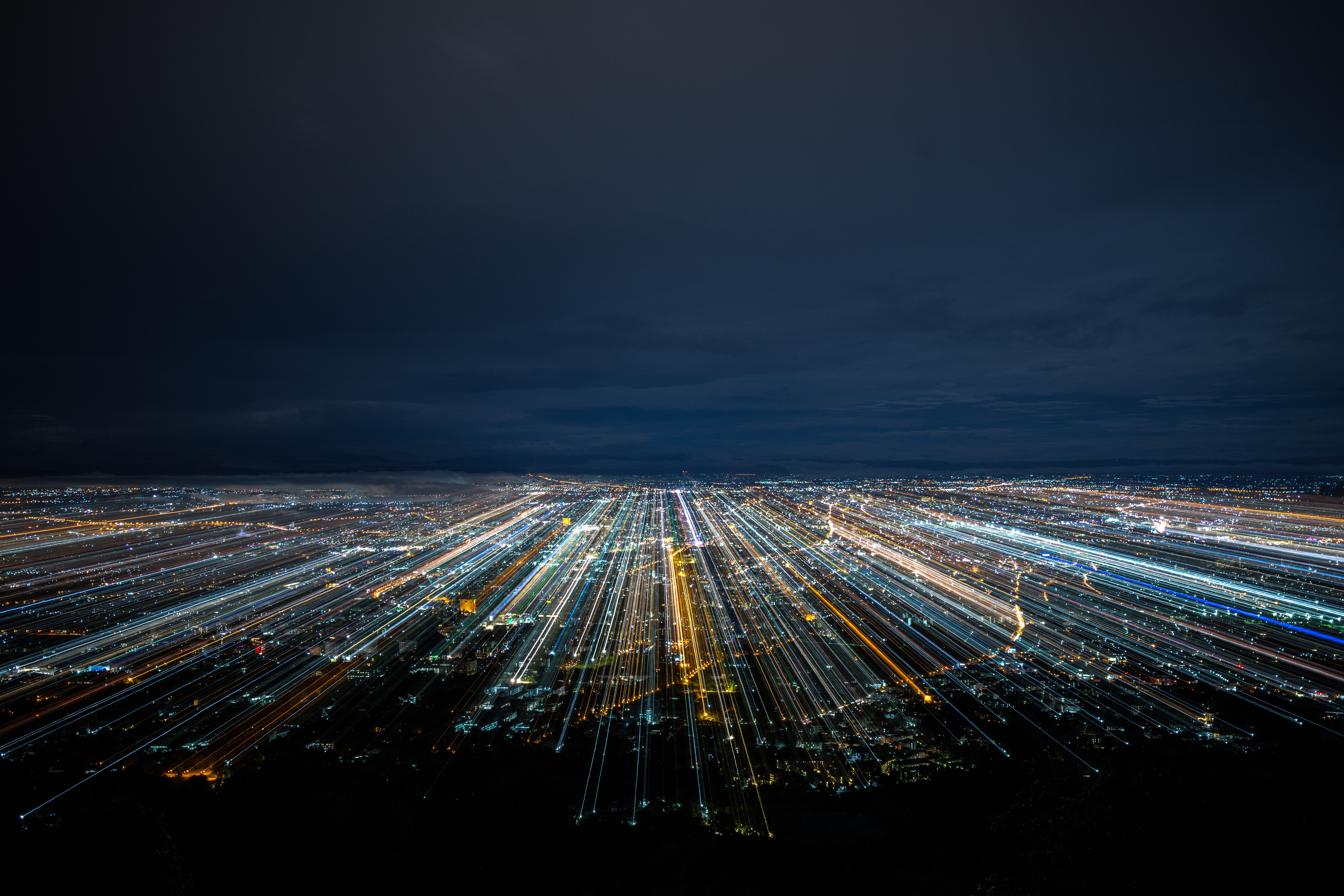 Aerial long-exposure photograph of a city at night, with streaks of light tracing road and infrastructure networks across the urban landscape.