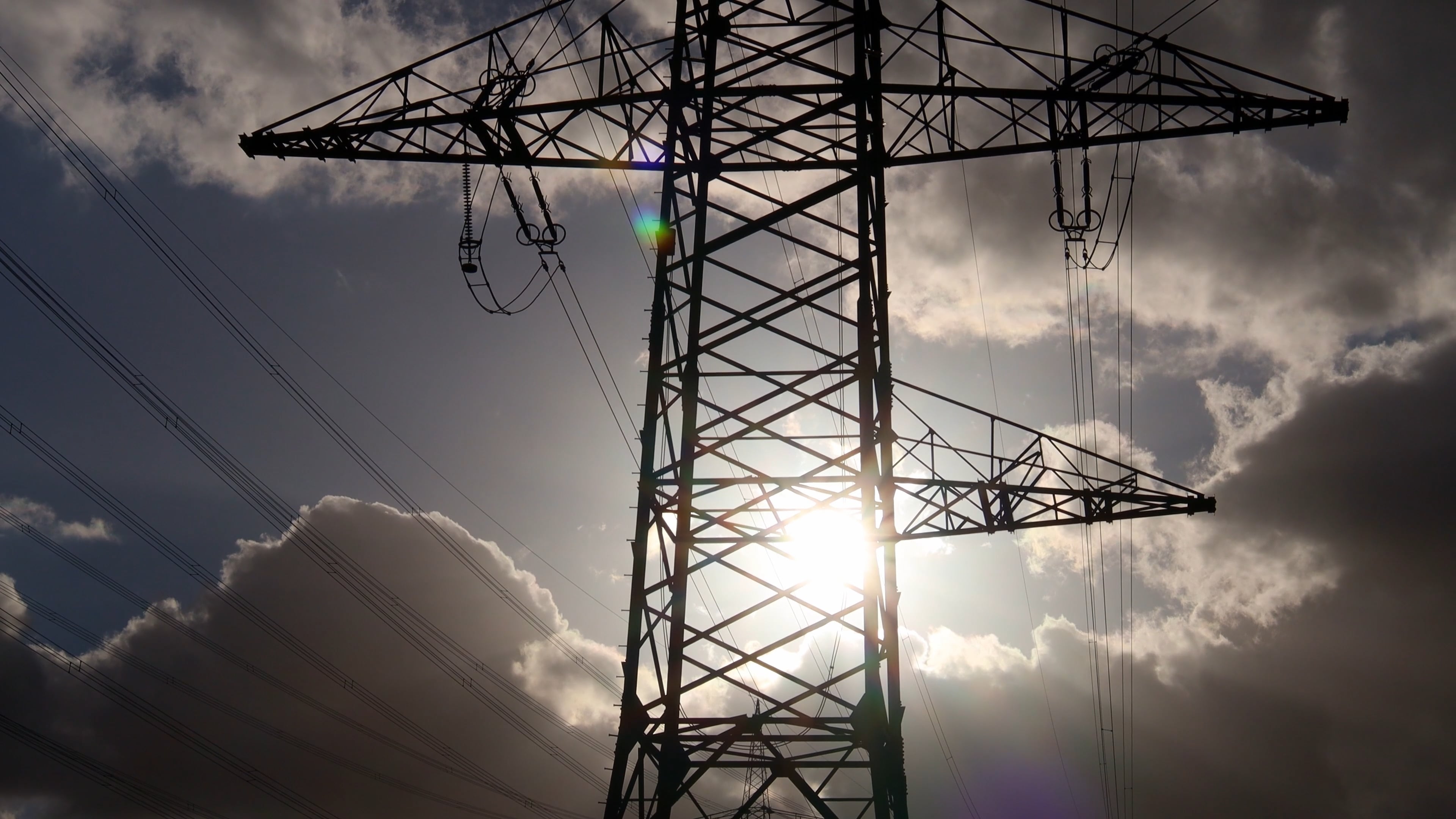 Silhouetted high-voltage transmission tower with multiple power lines against a dramatic sky as the sun shines through the structure.