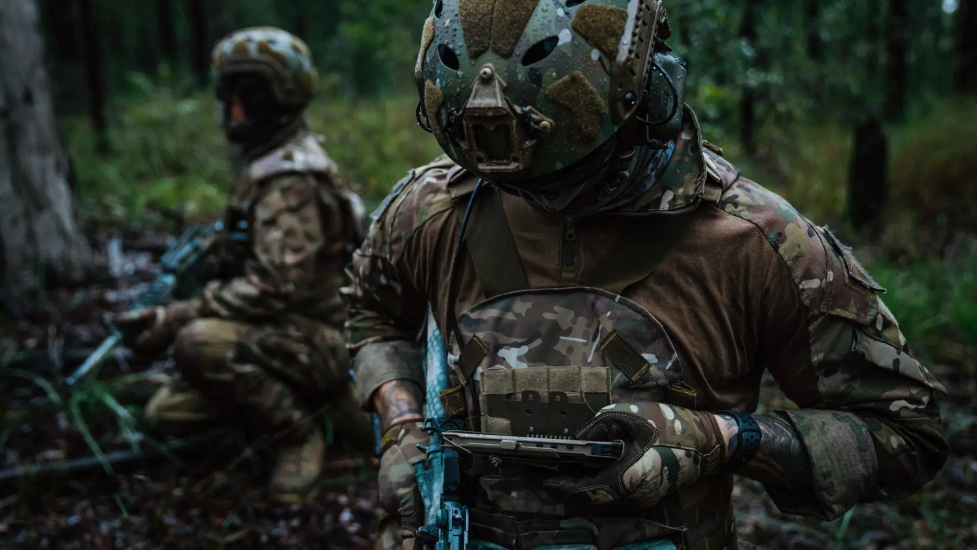 Two soldiers in camouflage gear use a handheld device with SitaWare Edge software during a tactical training mission in a wooded area.