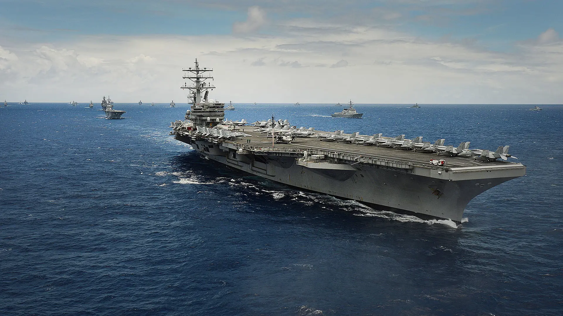 The USS Ronald Reagan aircraft carrier heads a fleet of warships in coordinated naval maneuvers on the open sea under a bright sky.