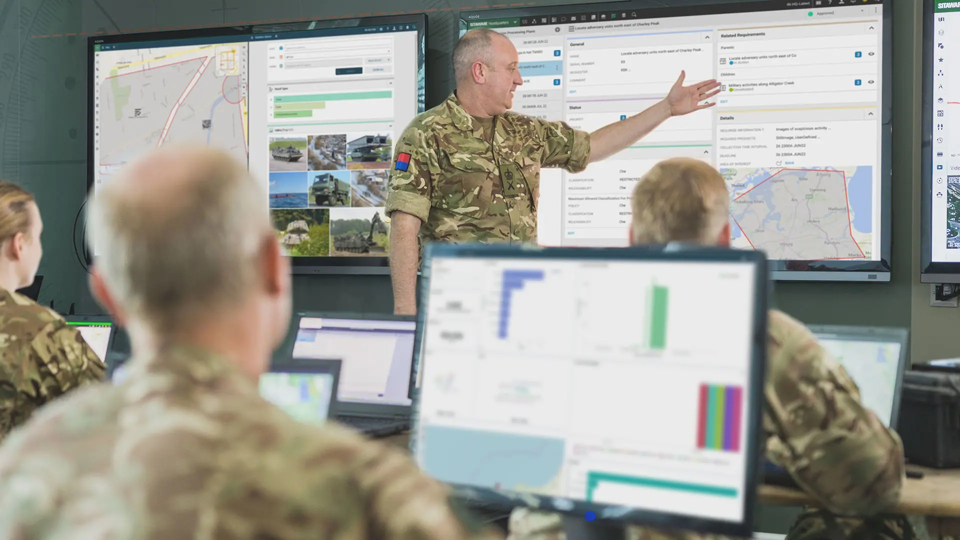 Soldiers in a command center analyze data as an officer presents on large screens.