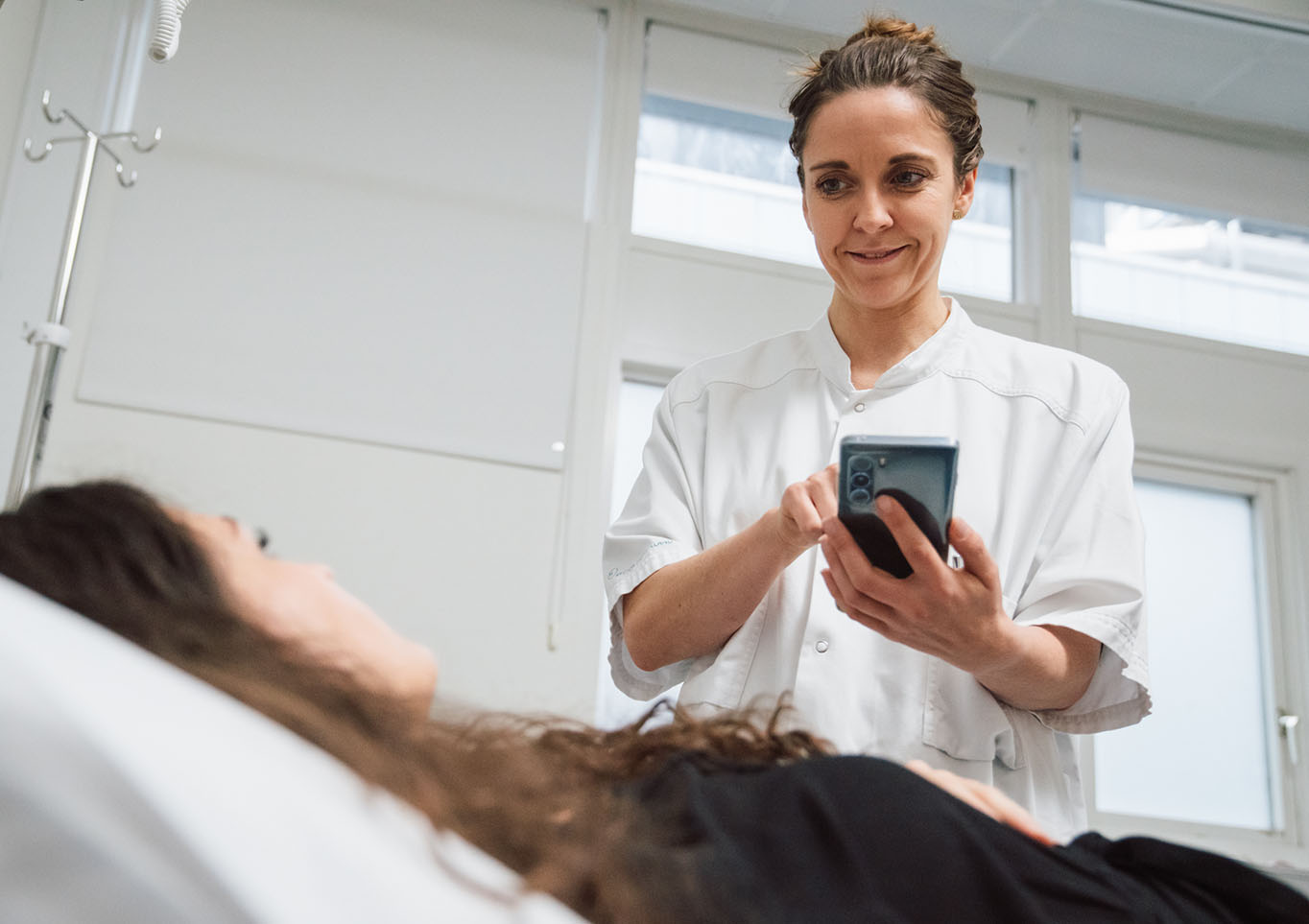 Nurse in clinical uniform using a smartphone at a patient’s bedside in a bright hospital room, illustrating digital documentation and modern patient care.