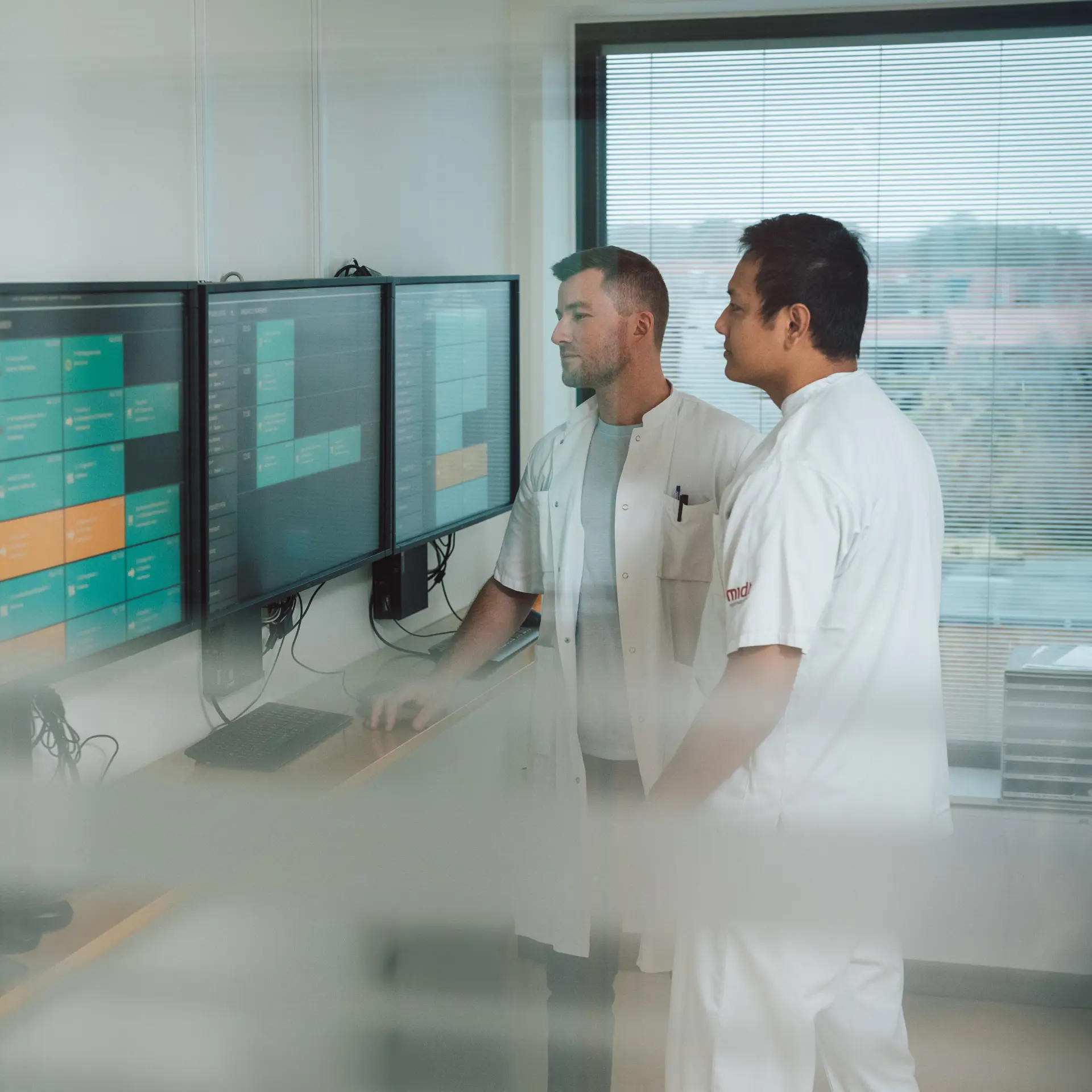 To sundhedsprofessionelle i hvide kitler analyserer data på flere store skærme i en medicinsk facilitet. Skærmene viser en struktureret tidsplan eller et patienthåndteringssystem.