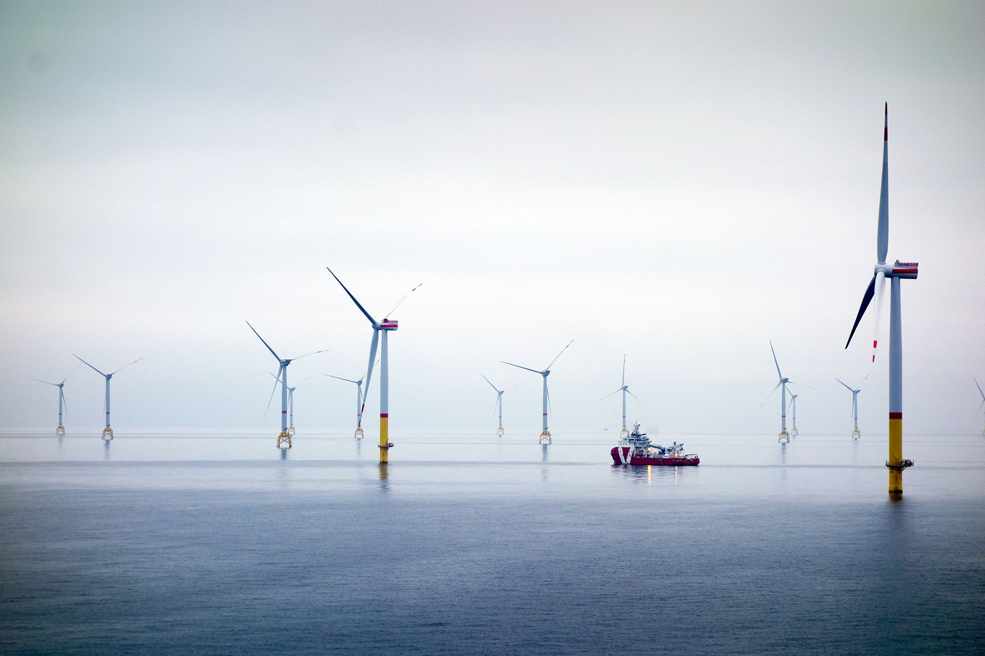 Offshore wind farm with multiple wind turbines and a service vessel operating at sea under overcast skies.