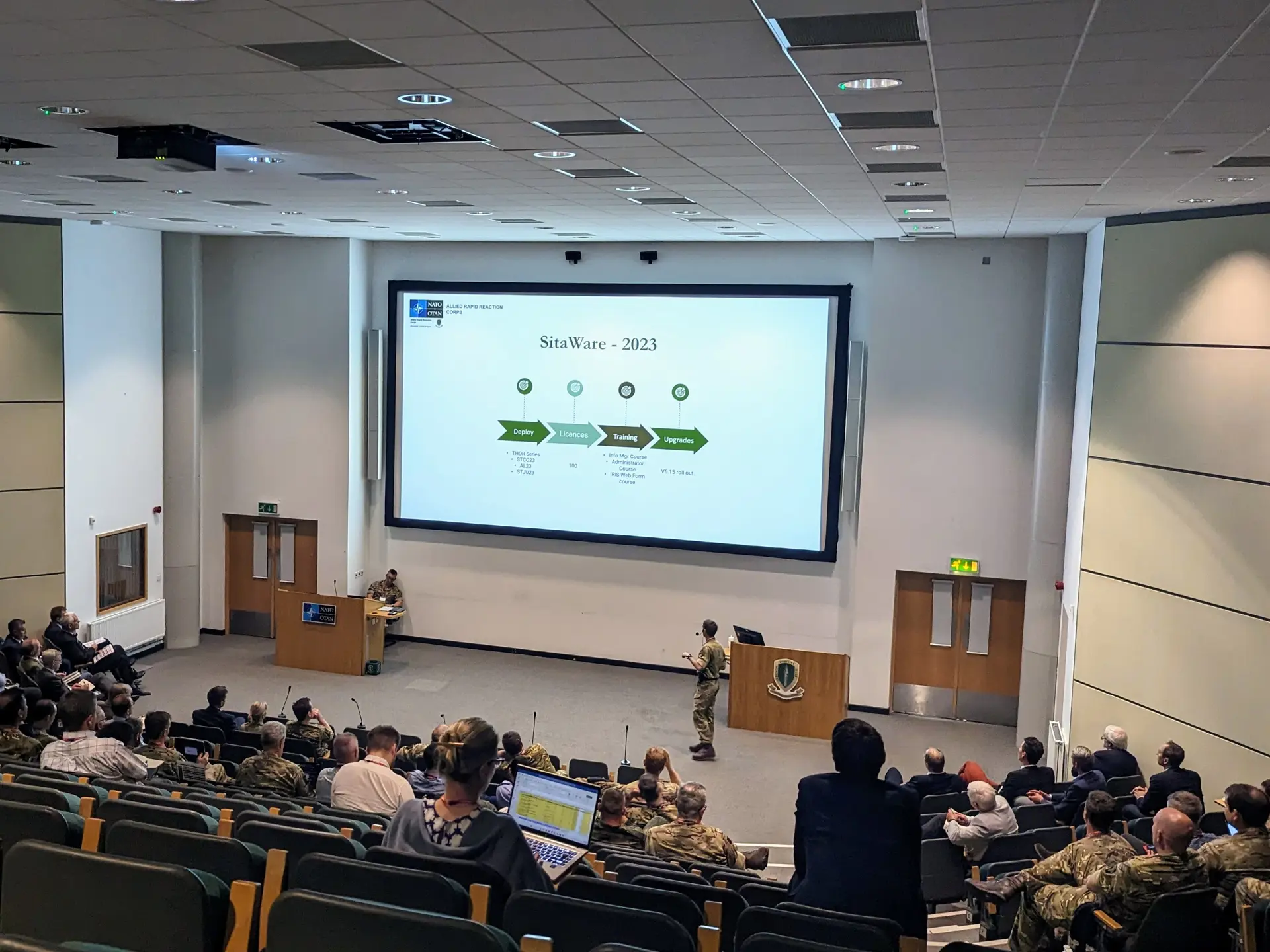Defense and military personnel gather at the SitaWare User Forum in the UK, engaging in a presentation in a spacious lecture hall.